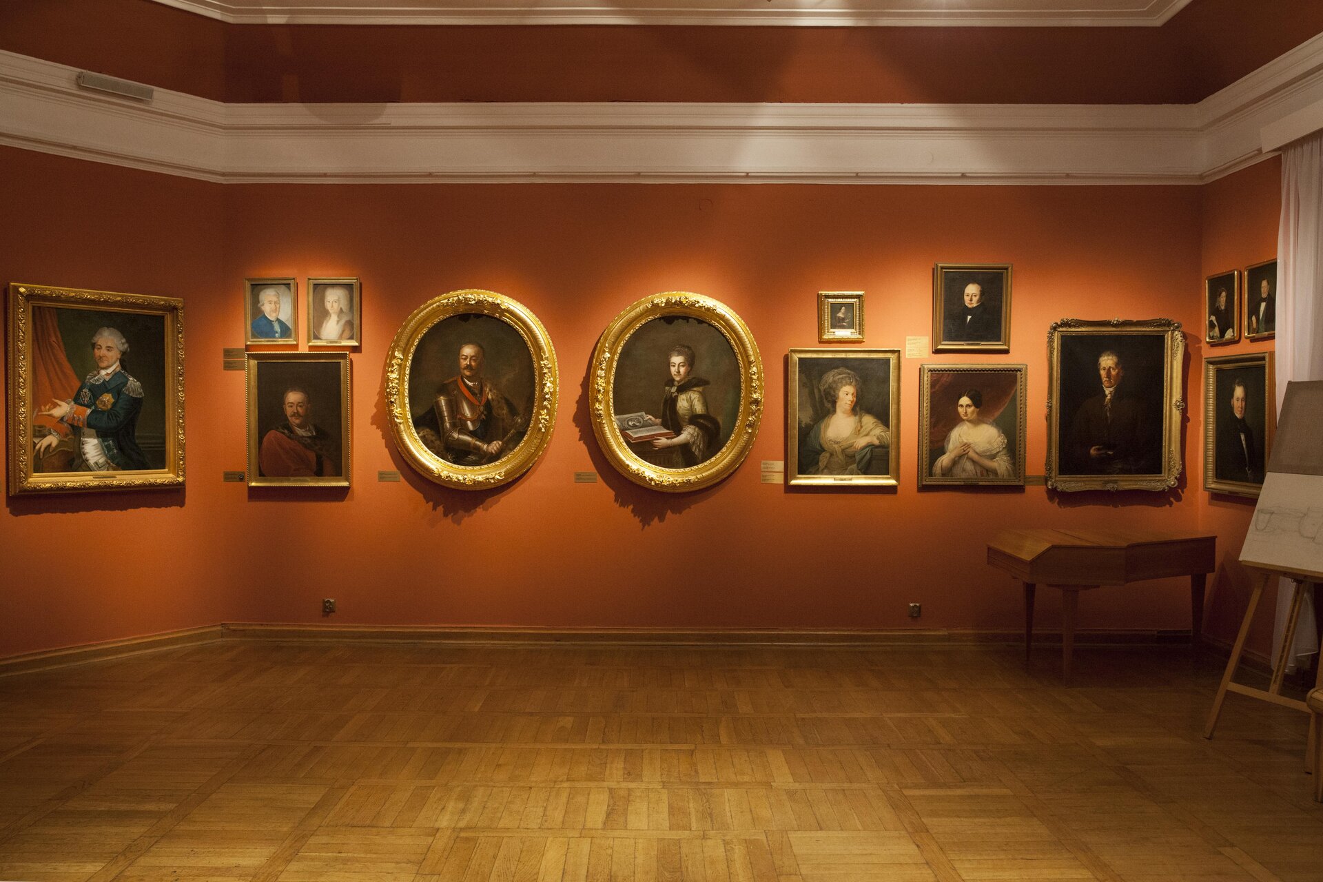 Zdjęcie ukazuje wnętrze galerii lub sali muzealnej gdzie prezentowana jest kolekcja dzieł sztuki. Na fotografii ukazana jest pomarańczowa ściana, na której wiszą obrazy. Obrazy mają różne kształty: kwadratowe, prostokątne oraz owalne. Wszystkie wykończone są złotą ramą. 