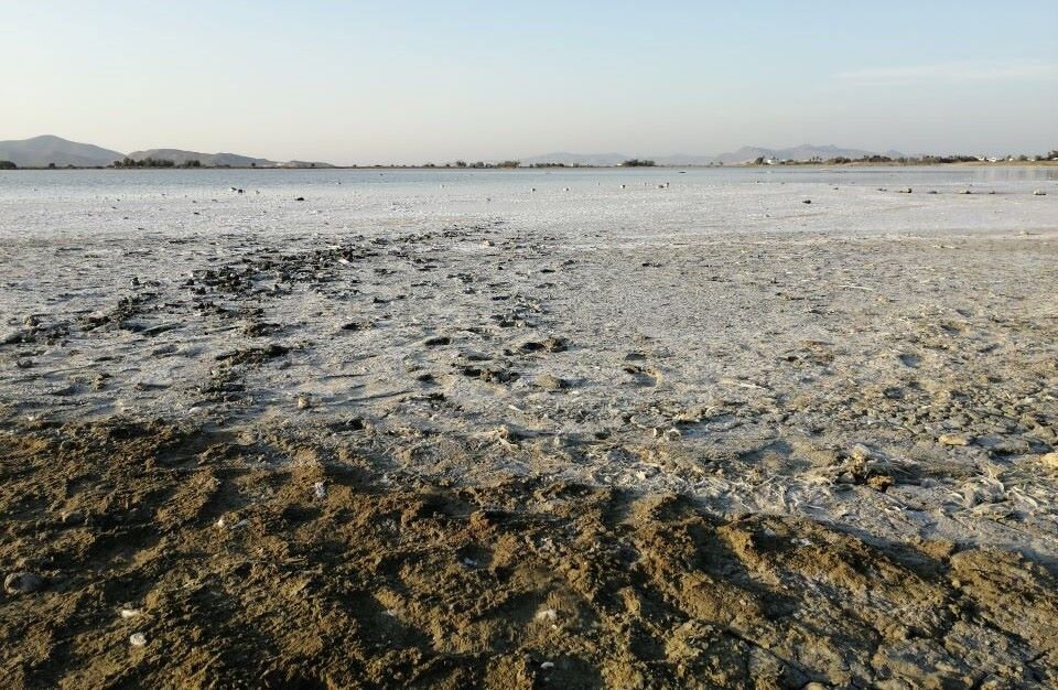 Ilustracja przedstawia fotografię, an którym znajduje się wysychające jezioro. W kadrze znajduje się rozległa panorama. Im dalej od planu pierwszego z wyraźnymi grudami piachu - krajobraz staje się coraz bardziej jednolity i coraz bardziej jaśniejszy.