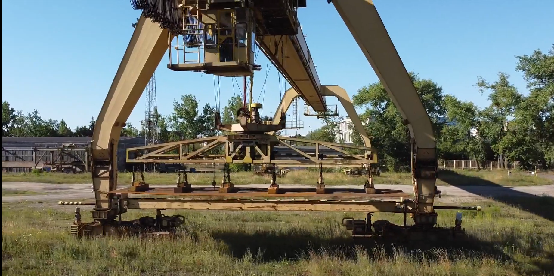 Zdjęcie przedstawia trawersę elektromagnetyczną. Trawersa jest podczepiona do suwnicy. Jest to duże urządzenie składające się z dwóch umieszczonych równolegle do siebie podstaw, które mają kształt odwróconej litery V. Połączone są ze sobą podłużnym ramieniem, na którym znajduje się wyciągnik z trawersą. Trawersa posiada liczne zakończenia z elektromagnesami, które są skierowane w dół. Przylega do nich transportowana blacha.