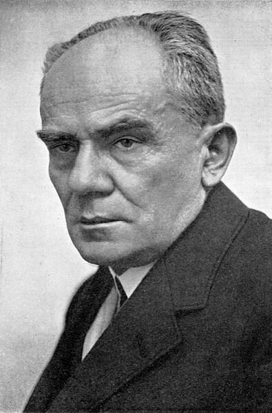 Czarno‑biała portretowa fotografia mężczyzny. Ma wysokie czoło, właściwie łysinę sięgającą do czubka głowy. Ma poważny wyraz twarzy. Ubrany jest w ciemną marynarkę, jasną koszulę.