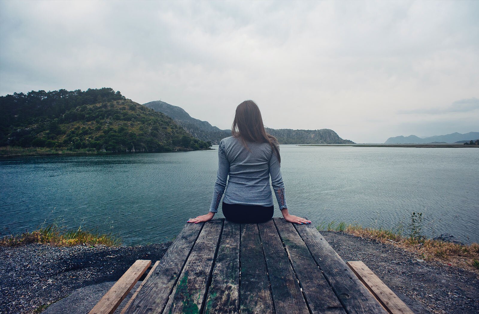 Zdjęcie przedstawia kobietę siedząca tyłem do obiektywu na stole z ławkami nad zbiornikiem wodnym. Widoczne są plecy kobiety ubranej w niebieską bluzkę, która patrzy przed siebie. Ma wyprostowane ręce, a otwartymi dłońmi dotyka stołu. W tle widoczne są porośnięte drzewami wzgórza, które otaczają zbiornik wodny.