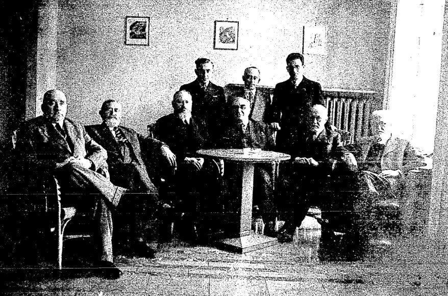 Zdjęcie przedstawia dziewięciu mężczyzn w pokoju. Na środku stoi niewielki okrągły stół wokół niego na fotelach siedzi sześciu mężczyzn, trzech stoi za za nimi. Wszyscy mają twarze zwrócone w kierunku obiektywu obiektywu. W tle znajduje się ściana na której wiszą 3 obrazki.
