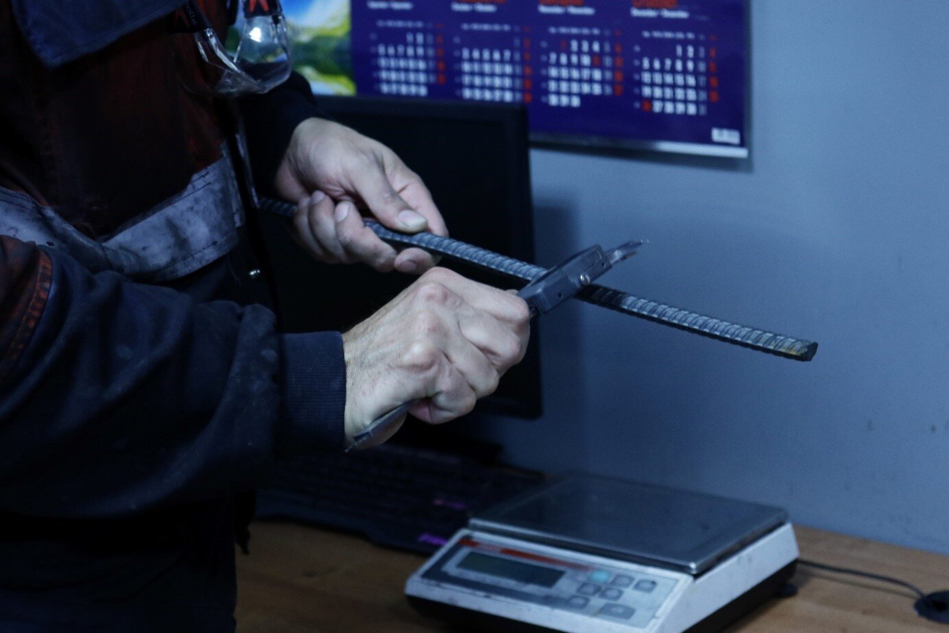 Zdjęcie przedstawia mężczyznę ubranego w strój pracowniczy dokonującego pomiarów geometrii i masy pręta walcowanego. Mężczyzna w jednej ręce trzyma pręt, a w drugiej suwmiarkę, którą dokonuje pomiaru średnicy pręta. Na biurku obok widoczna jest waga oraz monitor. W tle na ścianie wisi kalendarz.
