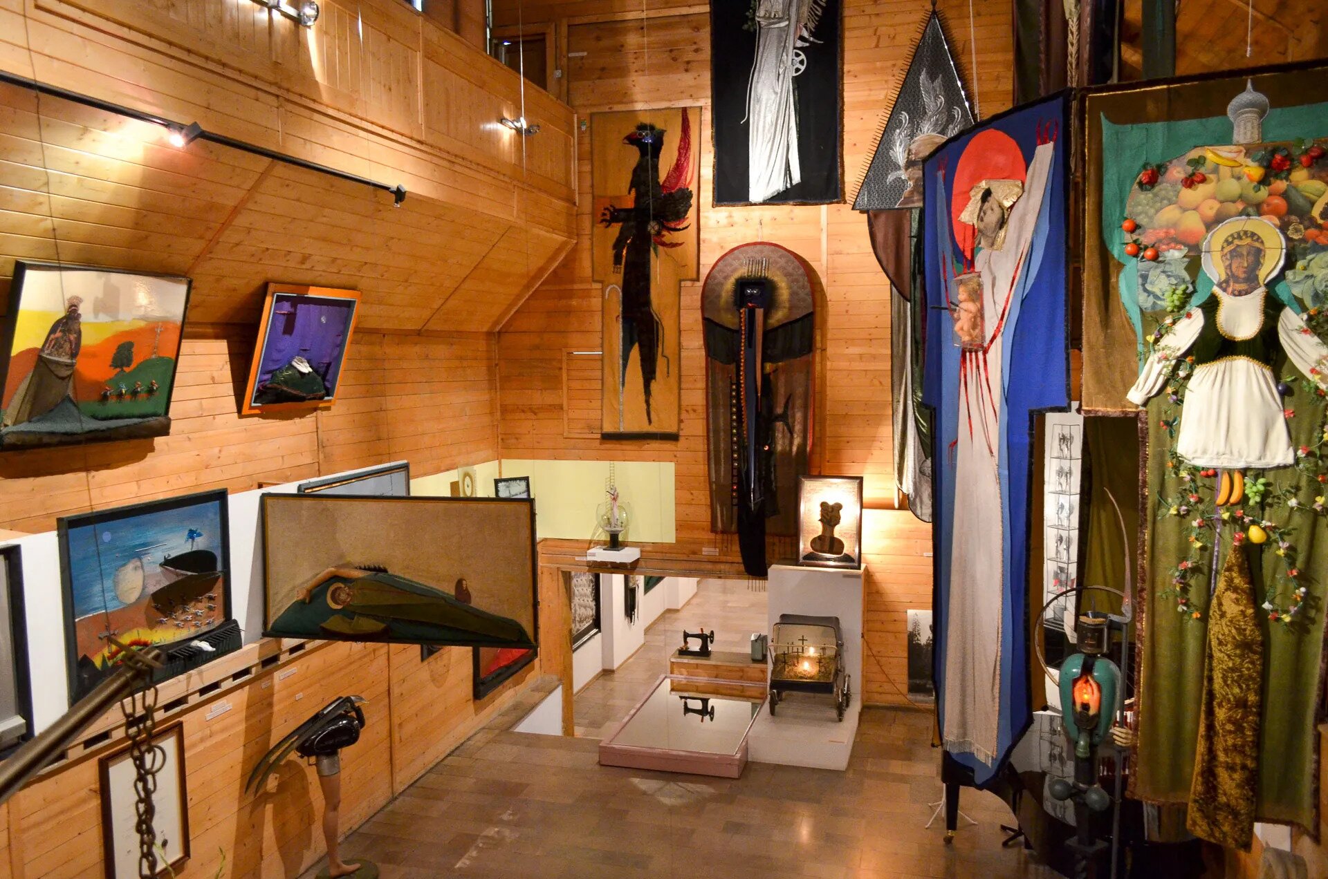 Zdjęcie przedstawia wnętrze galerii artystycznej. Na ścianach wiszą obrazy. Z sufitu zwisają dzieła na płótnie. W pracach przewijają się motywy religijne, przedstawienia postaci w aureolach, jedna otoczona owocami, druga  zdrewnianą, trójwymiarową ręką. Na ścianie wisi wizerunek istoty podobnej do czarnego ptaka. Dzieła składają się z różnych materiałów i mają nieregularne kształty.