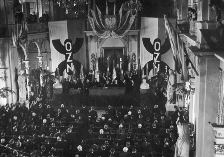 Zdjęcie przedstawia salę zapełnioną krzesłami, na których siedzą zgromadzeni mężczyźni. W centralnej części znajduje się stół, za którym siedzą mężczyźni prowadzący spotkanie. Nad nimi umieszczone jest godło Polski oraz sztandary z napisem OZN. W sali widoczne są kryształowe, zdobione żyrandole i łuki kolumnowe. 