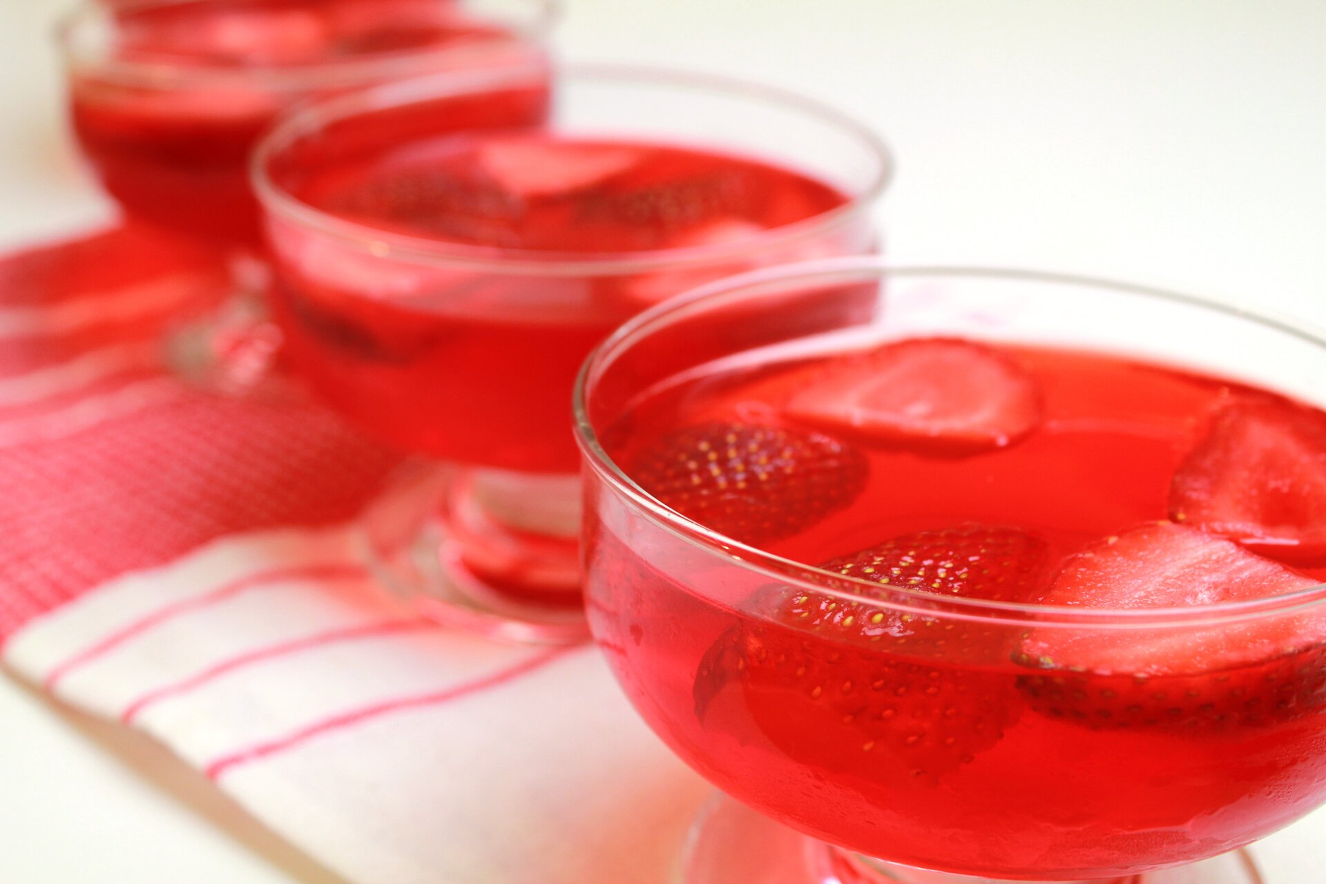 Zdjęcie przedstawia trzy salaterki ze słodką galaretką, w której zatopiono połówki truskawek. Salaterki stoją na białej ściereczce w czerwony wzór. Ostra jest tylko pierwsza, pozostałe dwie znajdują się poza obszarem ostrości fotografii.