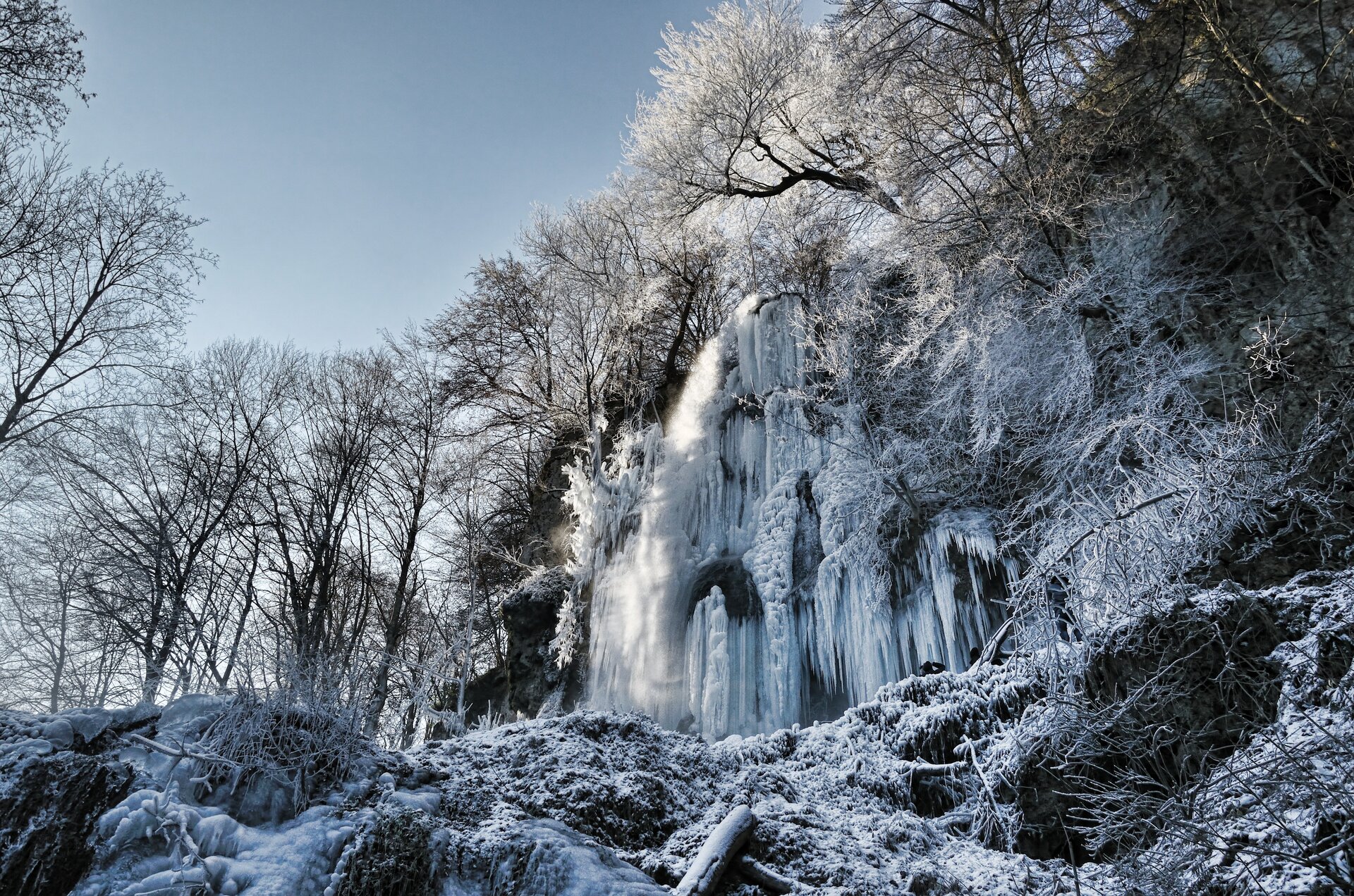 Zdjęcie przedstawia wodospad zimą uwieczniony od jego podstawy. Najbliższe okolice wodospadu oraz większa część jego powierzchni pokryte są lodem. Drzewa rosnące poniżej skalnej półki, z której wypływa woda również są silnie oszronione. Pozostałe drzewa mają nagie gałęzie. Prawa i dolna krawędź kadru zajęta przez ośnieżone skały. W lewym górnym rogu kadru niebieskie bezchmurne niebo.