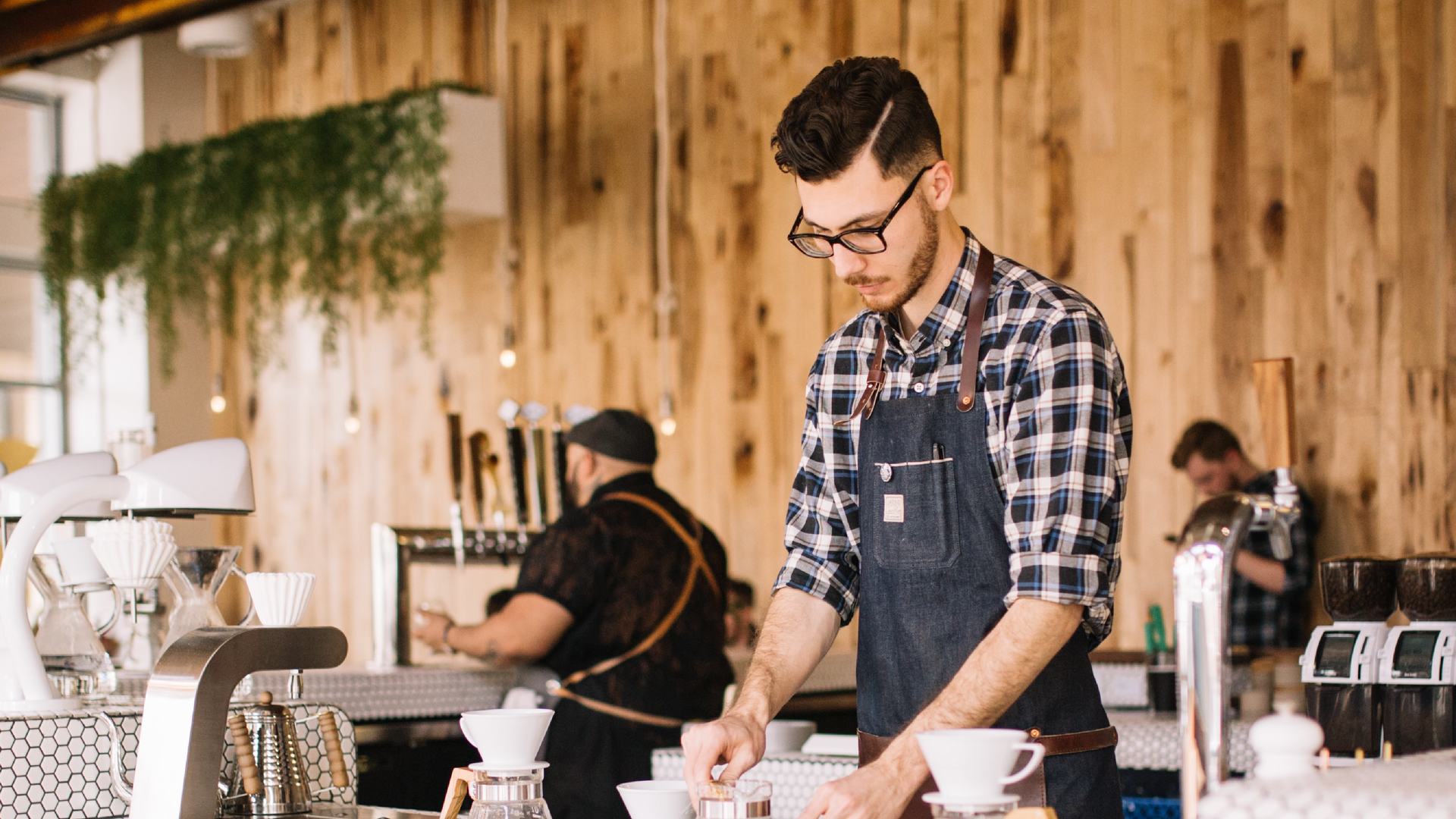 Zdjęcie przedstawia młodego wysokiego mężczyznę w barze. Mężczyzna nosi okulary, koszulę w kratkę i fartuszek zakładany przez głowę. Mężczyzna przygotowuje filiżanki na kawę. W tle widoczny jest inny mężczyzna, który przygotowuje kawę w dużym ekspresie. Mężczyzna stoi tyłem do kadru.