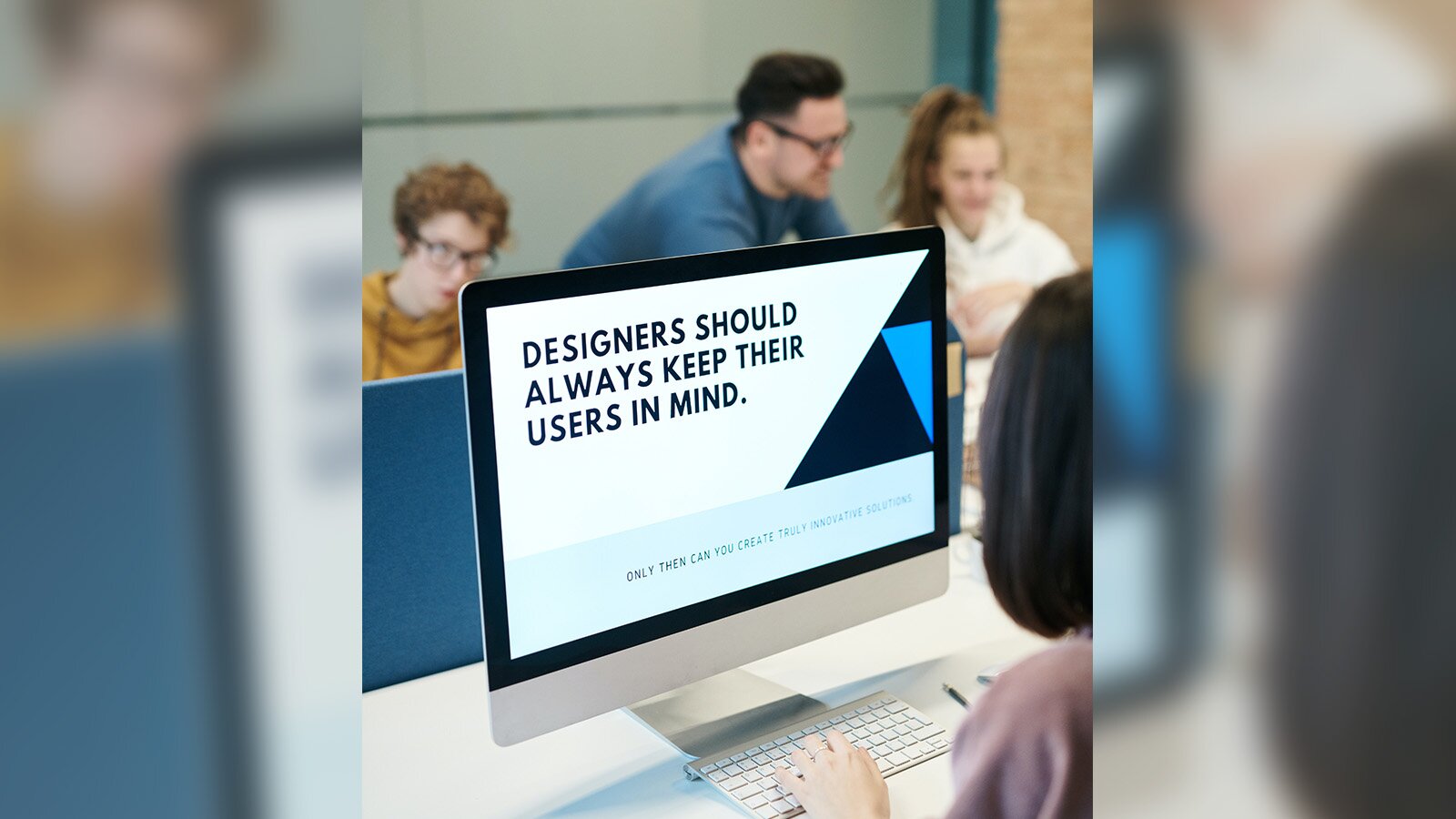 Zdjęcie przedstawia osobę przed ekranem komputera, na którym ukazano napis "DESIGNERS SHOULD ALWAYS KEEP THEIR USERS IN MIND". Za monitorem widoczne trzy osoby, mężczyznę w średnim wieku, nastolatkę i chłopca w wieku około 10 lat.
