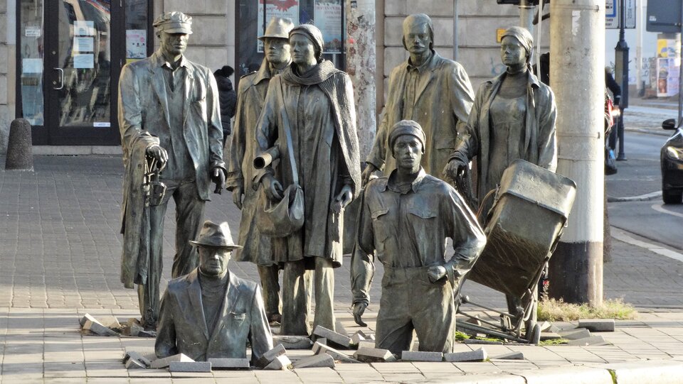 Zdjęcie przedstawia pomnik autorstwa Jerzego Kaliny, pod tytułem „Pomnik anonimowego przechodnia”. Dzieło przedstawia ludzi, postacie z brązu naturalnej wielkości. Na fotografii ukazanych jest siedem osób. Na pierwszym planie, na środku znajdują się dwaj mężczyźni, którzy znikają w chodnikowych płytach. To sprawia, że tworzy się iluzja zapadającej się ziemi.  Za mężczyznami  po prawej stronie grupy figur można wyróżnić kobietę z dziecięcym wózkiem oraz mężczyznę z walizką. W środku pomiędzy mężczyznami  znajduje się kobieta z torebką oraz zwojem papieru. Po lewej stronie za mężczyzną  znikającym w płytkach stoi starszy mężczyzna z laską, a tuż obok niego mężczyzna w kapeluszu. Cała kompozycja figur znajduje się na chodniku, tuż przy ulicy. W oddali widać budynki.  