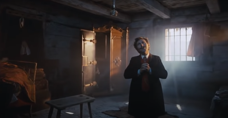 Głównym bohaterem tego filmowego kadru jest klęczący mężczyzna pogrążony w modlitwie. Jego głowa uniesiona jest ku górze, a splecione dłonie spoczywają na piersi w geście skupienia i oddania. Jasne, kręcone włosy oraz wąs pod nosem nadają jego twarzy wyraz powagi i emocjonalnego napięcia. Ubrany jest w ciemny surdut, biały wykrochmalony kołnierz koszuli oraz ciemny krawat, co podkreśla uroczysty charakter chwili.

Scena rozgrywa się w lekko przyciemnionej izbie, której klimat budują proste, nieco surowe meble. Po lewej stronie widać metalowe łóżko, obok którego stoi duża, otwarta szafa. W centralnej części pomieszczenia, tuż przy mężczyźnie, znajduje się masywny taboret. Nad nim, przy suficie ozdobionym drewnianymi belkami, wisi stara lampa naftowa, rzucająca ciepłe, przygaszone światło na modlitewną scenę.