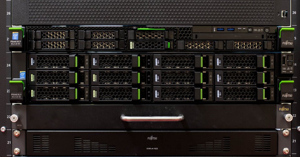 Obraz przedstawia serwer zamontowany w szafie rackowej. Widzimy przedni panel urządzenia z wieloma kieszeniami na dyski twarde, z których część ma zamontowane dyski. Każda kieszeń jest zabezpieczona kratką wentylacyjną z zielonymi uchwytami do szybkiego demontażu. Po lewej stronie znajdują się oznaczenia modeli serwerów oraz naklejki informujące o procesorach. W środkowej części znajdują się porty USB i przyciski zasilania/resetu. Na dole widoczny jest większy panel z uchwytem oraz logo producenta. 