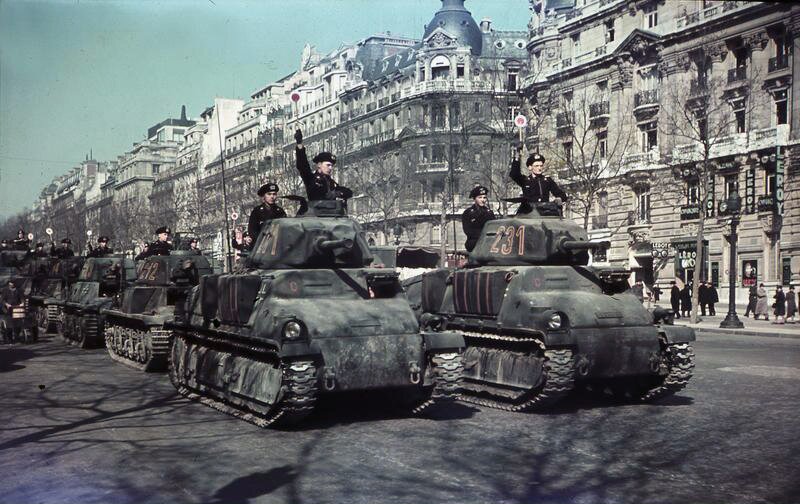 Zdjęcie przedstawia niemieckie lekkie czołgi jadące ulicą. Z ich wieżyczek wychylają się niemieccy żołnierze, którzy wyciągają przed siebie ręce w hitlerowskim pozdrowieniu. W tle widać wysokie kamienice oraz kilkoro ludzi spacerujących chodnikiem.