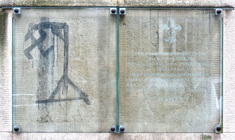 Na zdjęciu znajduje się fragment ściany budynku. Po lewej stronie czarną farbą została na niej narysowana swastyka powieszona na szubienicy.  Rysunek zasłonięty jest szkłem. Obok niego jest szklana tablica z tekstem.