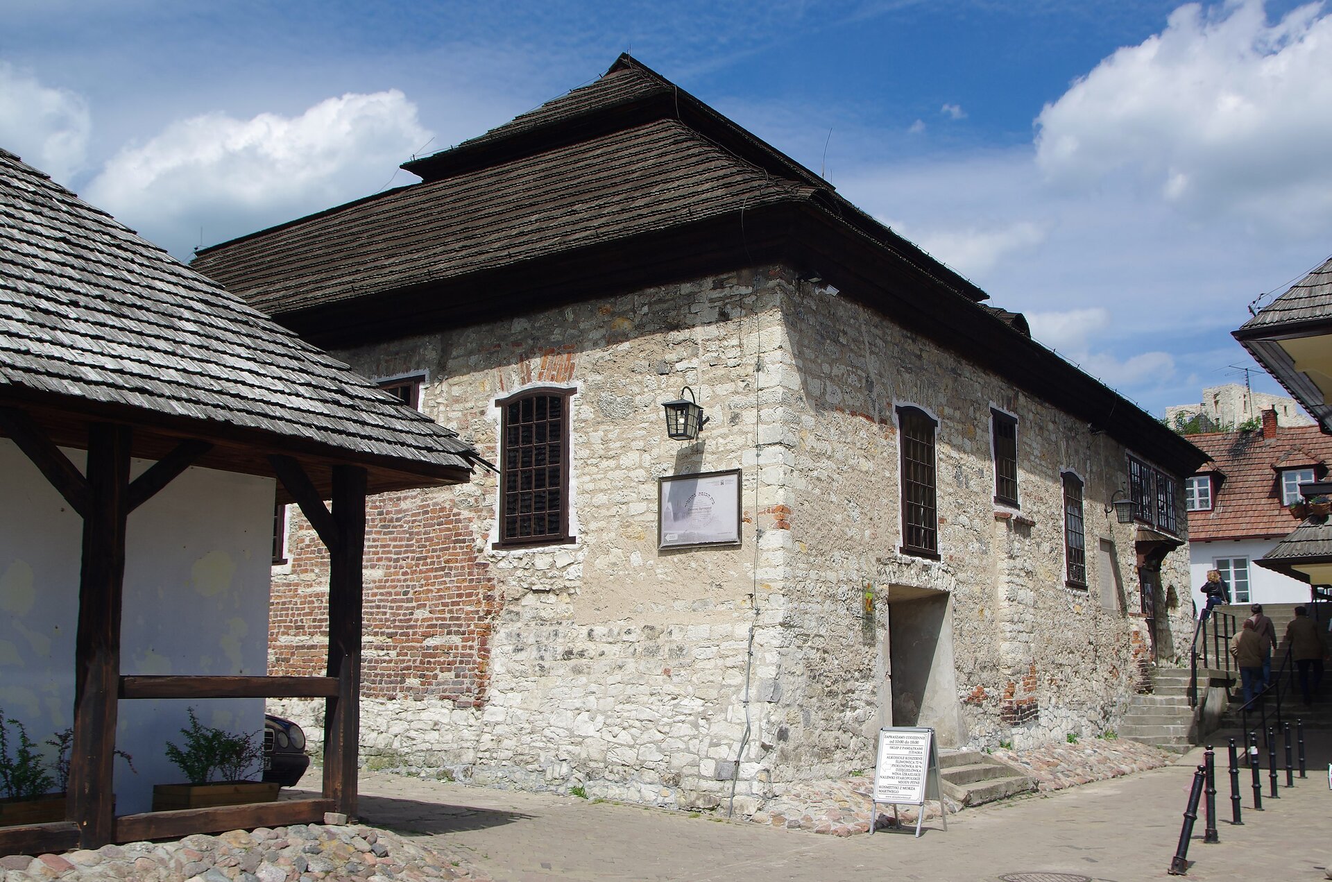 Na zdjęciu stary,  murowany budynek. Ma niewysoki, czarny dach i ściany z białej cegły. W białych ścianach budynku znajdują się brązowe okna. Dookoła budynku jest wybrukowana uliczka po której spacerują ludzie. Po lewej stronie fotografii widoczny jest róg innego budynku. 