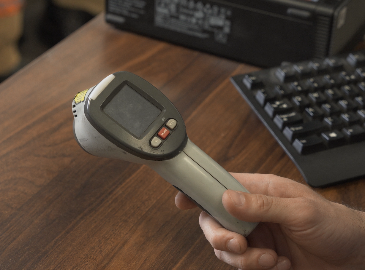 Zdjęcie przedstawia pirometr trzymany w dłoni. W tle znajduje się biurko, na nim klawiatura. Pirometr wyposażony jest w uchwyt pistoletowy. Na nim osadzona jest nieduża prostopadłościenna część odpowiadająca za pomiar. Po jednej stronie znajduje się prostokątny wyświetlacz. Poniżej ekranu wbudowane są trzy przyciski.