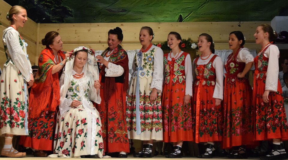 Zdjęcie przedstawia grupę kobiet w ludowych góralskich strojach. Dwie starsze kobiety zakładają chustę na głowę młodej kobiety. Reszta kobiet śpiewa.