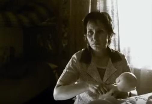 Zdjęcie przedstawia kobietę w jasnym fartuchu, która siedzi przy stoliku i trzyma lalkę. Patrzy przed siebie, ma niedługie, ciemne włosy. Za nią jest okno z firanką.