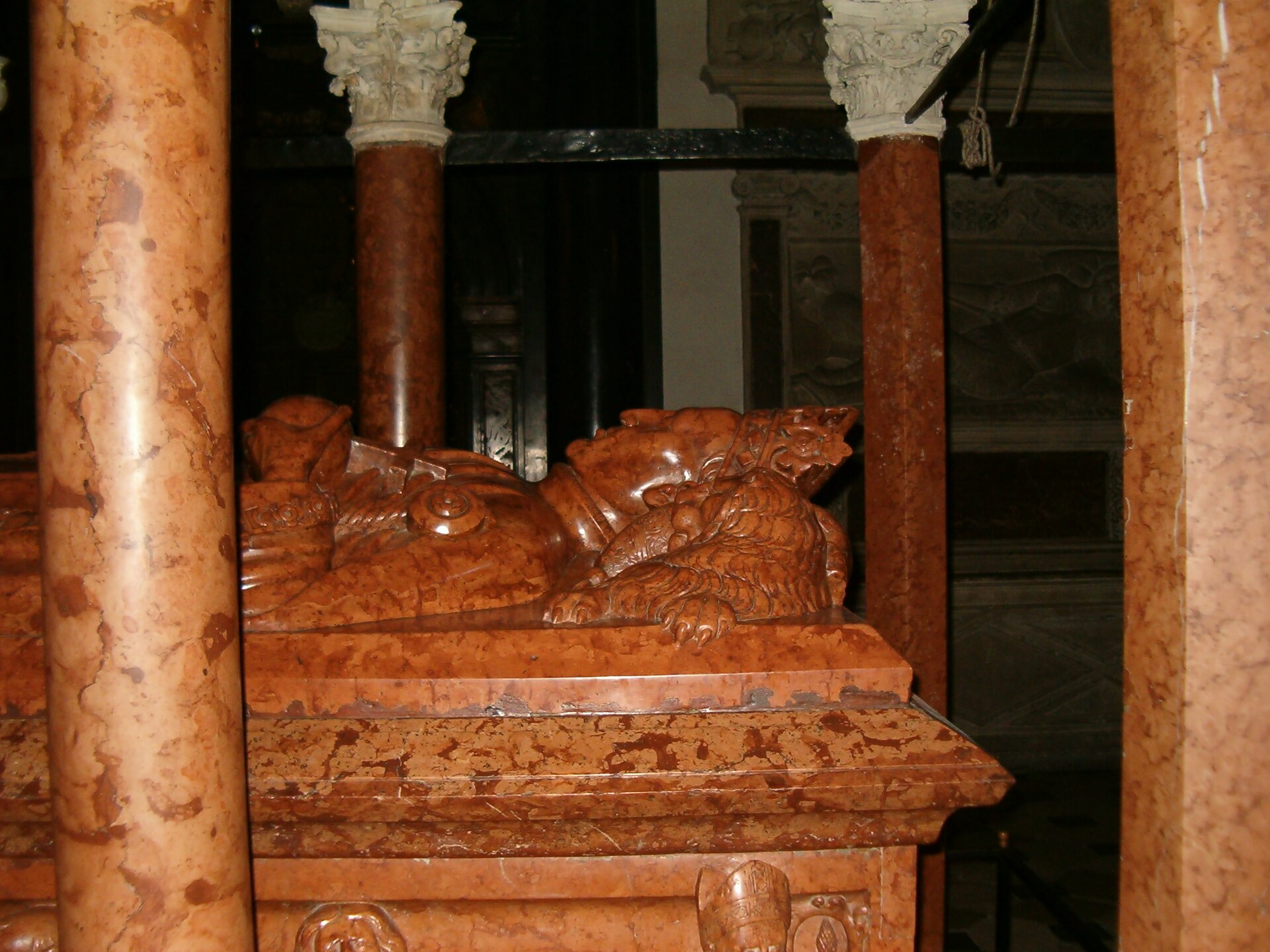 Zdjęcie przedstawia marmurowy nagrobek mężczyzny. Widzimy fragment sarkofagu, ma którym widać podobiznę zmarłego od głowy do piersi. Dalsza część rzeźby zasłonięta jest przez jedną z kolumn, które stoją wokół nagrobka. Zwieńczenie kolumn, tak zwane głowice są bogato rzeźbione w roślinny motyw. Postać mężczyzny ma głowę położona na podwyższeniu, na głowie ma koronę. W rękach trzyma berło i krzyż.  