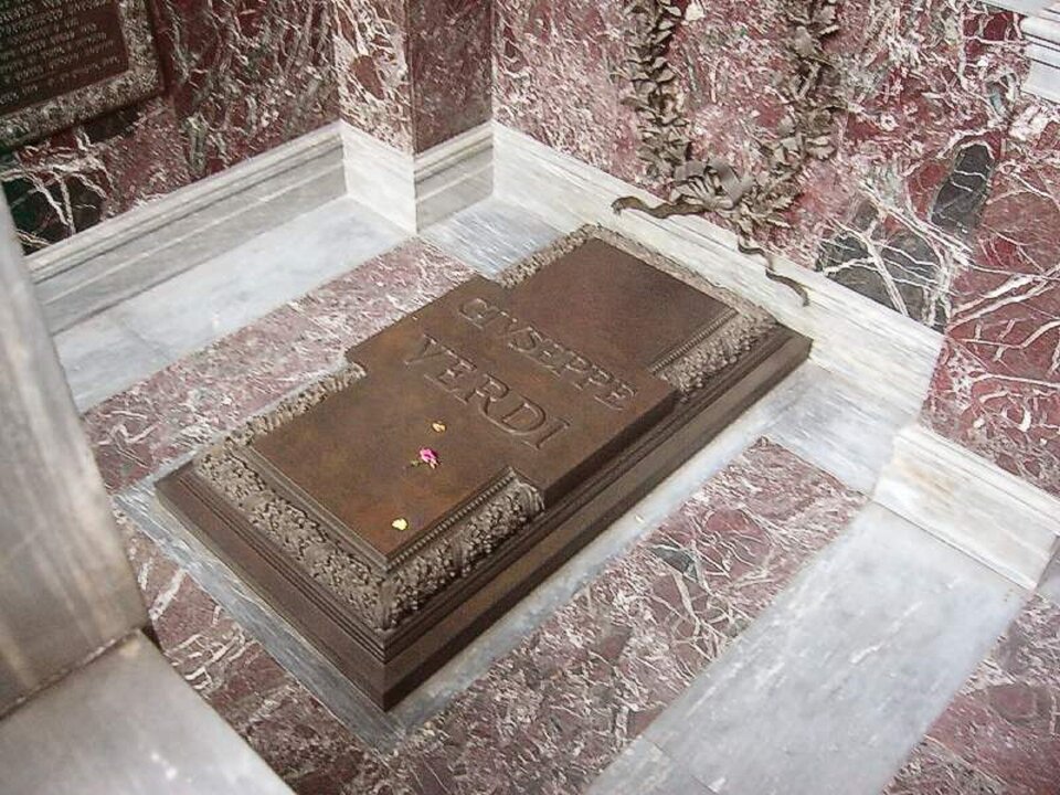 Ilustracja przedstawia: Grób Verdiego w Mediolanie. Fotografia przedstawia nagrobek z marmurowej płyty ze zdobieniami na jej brzegach. Na środku widoczny jest napis GIUSEPPE VERDI.