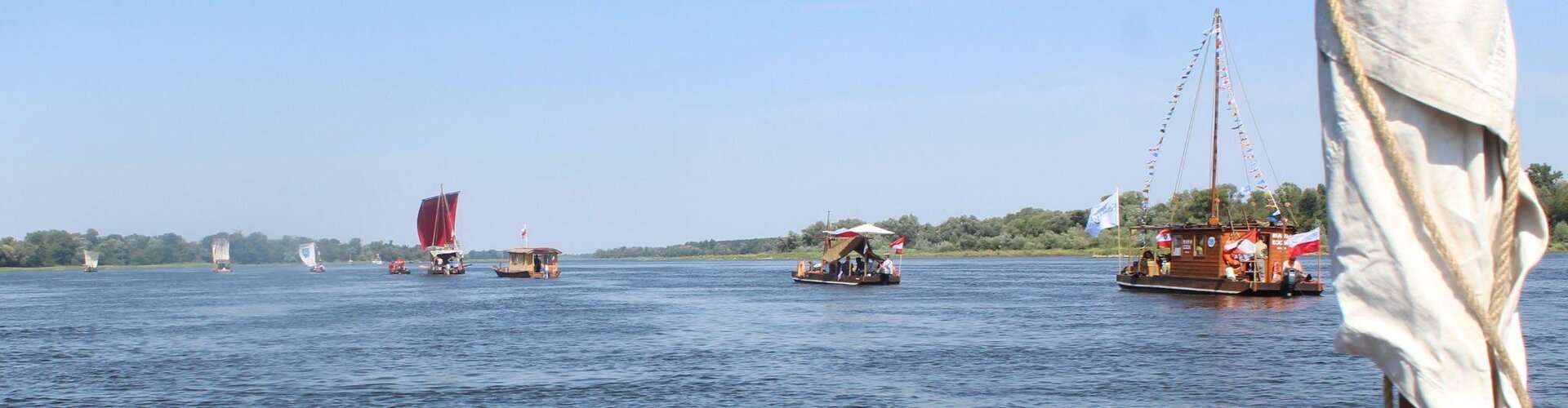 Zdjęcie przedstawia panoramę rzeki, na której znajdują się żaglówki. Po prawej stronie widać mały statek turystyczny, przystrojony kolorowymi flagami. Przed nim, w pewnej odległości, płynie łódka z białym, materiałowym dachem. W centralnej części rzeki, w oddali, widoczny jest jeszcze jeden drewniany statek wycieczkowy, a także żaglówka z czerwonym żaglem, a przed nią trzy inne, z białymi żaglami. Z prawej strony zdjęcia, na pierwszym planie, znajduje się fragment białego żagla przewiązanego jasnobrązowym sznurkiem. Rzeka jest spokojna, błękitna. Przy jej obu brzegach widoczna jest bujna, zielona roślinność. Niebo jest błękitne, bezchmurne. Zdjęcie zostało zrobione w świetle dziennym.