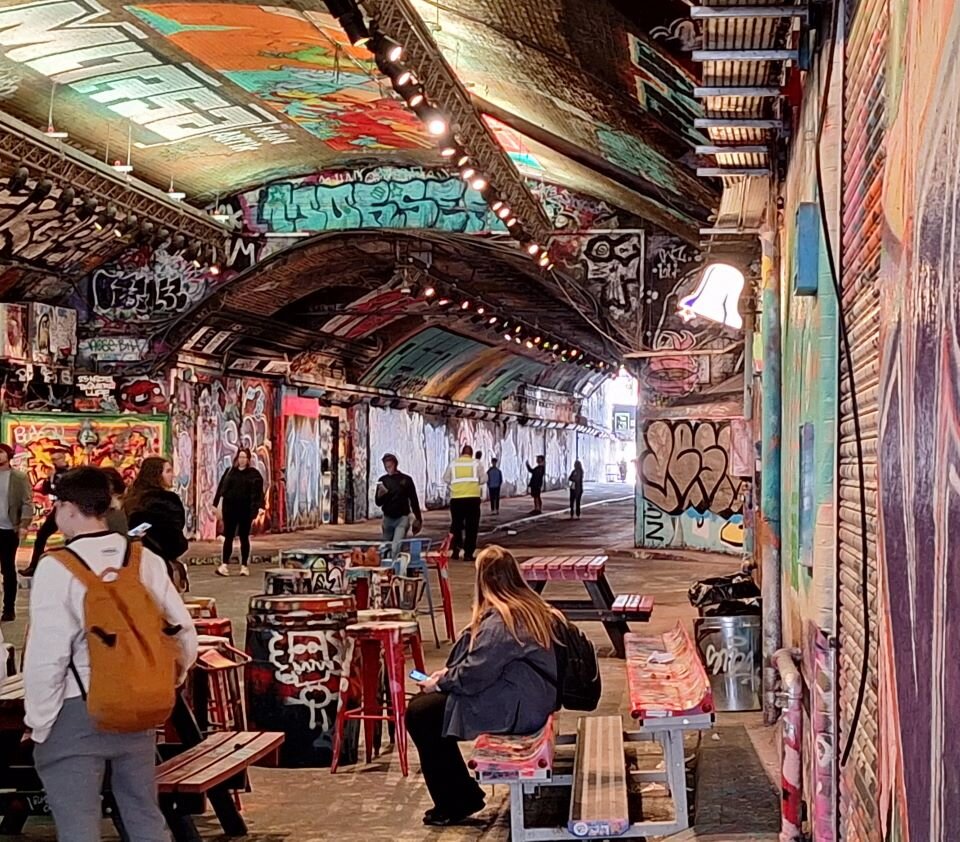 Ilustracja przedstawia fragment tunelu pokrytego malowidłami. Graffiti jest wielokolorowe. W tunelu znajduje się wiele ludzi: przechodzących przez tunel, siedzących pod ścianami tunelu oraz oglądających malowidła malowidła.