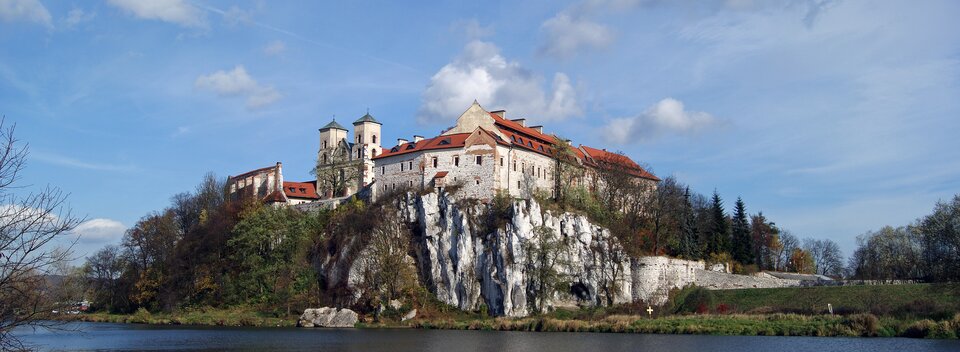 Na zdjęciu znajduje się klasztor w Tyńcu. Ma on kamienne ściany i czerwony dach. W jego centralnej części znajdują się dwie niskie wieże. Jest on umiejscowiony na lekkim kamiennym wzniesieniu o stromych zboczach. Przed nim płynie rzeka. 