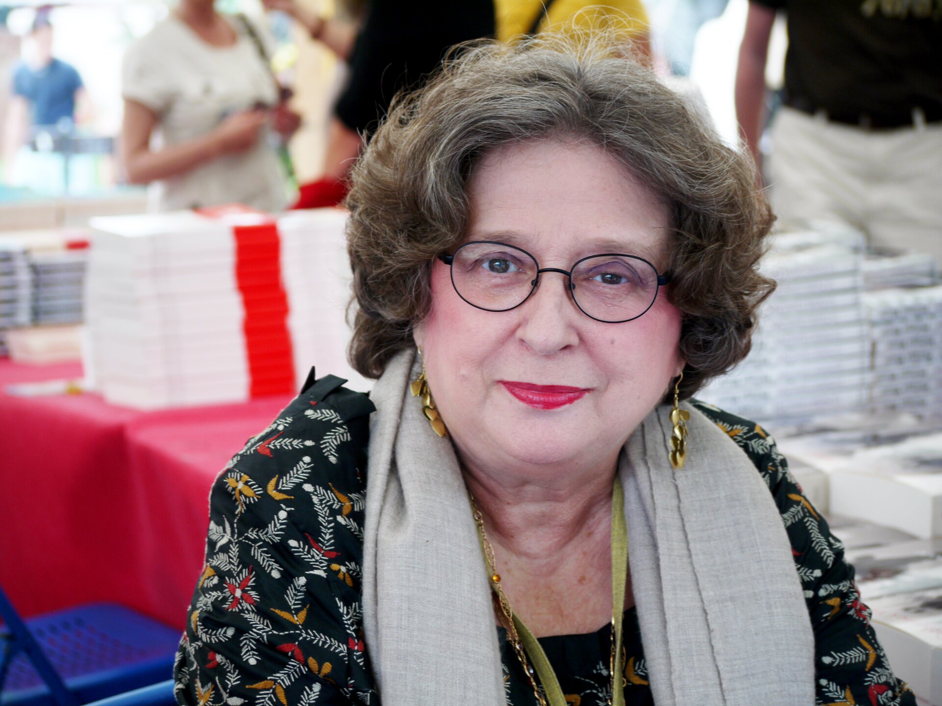 Na zdjęciu znajduje się starsza kobieta w okularach o okrągłej twarzy i kręconych włosach. Usta ma pomalowane wiśniową szminką. W uszach ma długie kolczyki. Lekko się uśmiecha. Na szyi ma zawieszony szary szal. W tle poukładane w stosy książki.