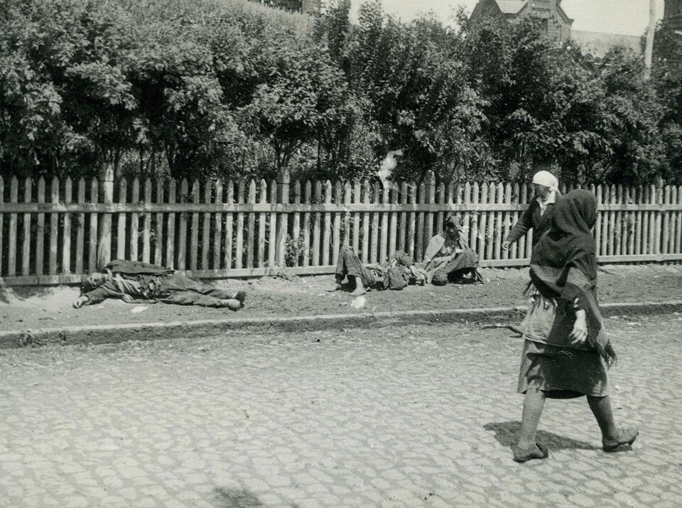 Zdjęcie przedstawia drewniany płot, pod którym leżą martwe, wychudzone ciała. Mijane są przez dwie kobiety w prostych sukniach z chustami na głowach, które idą ulicą. Jedna z nich spogląda na ciała. W tle rosną drzewa.
