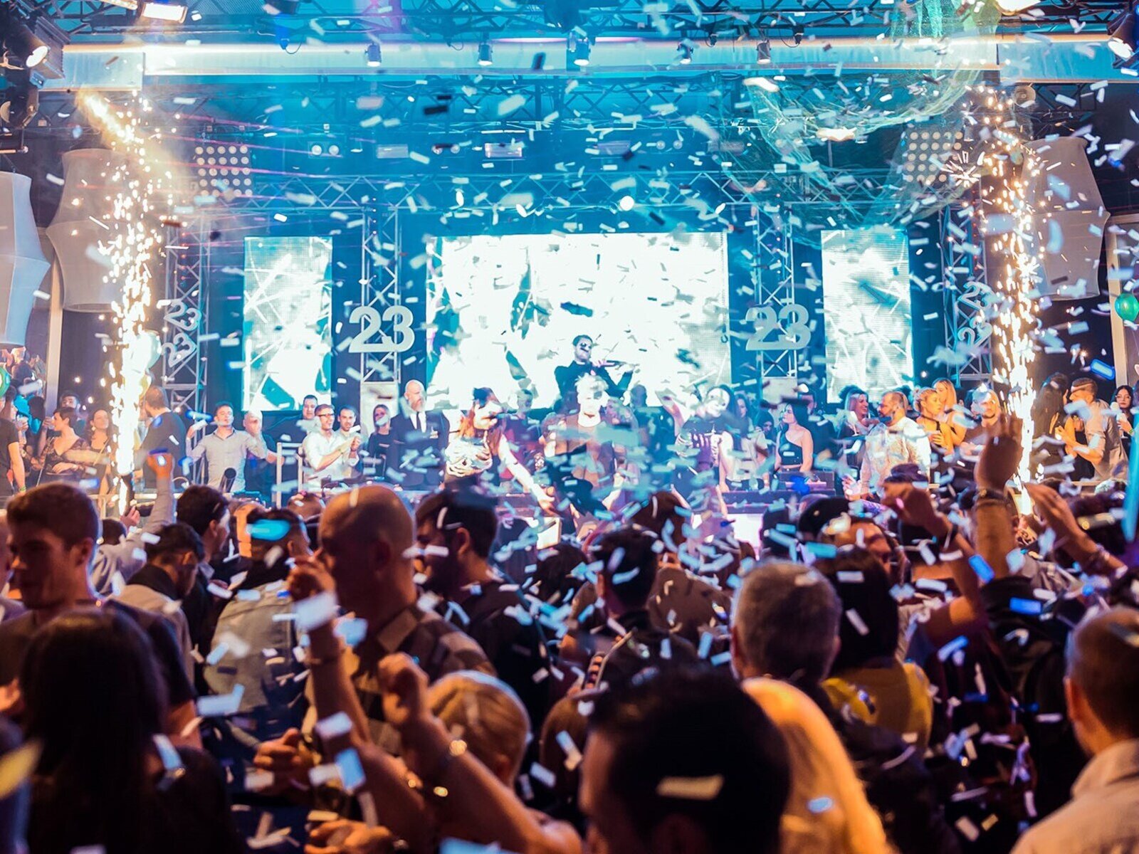 Zdjęcie przedstawia  tłum ludzi przed sceną niektórzy mają ręce w górze, z sufitu spada confetti.