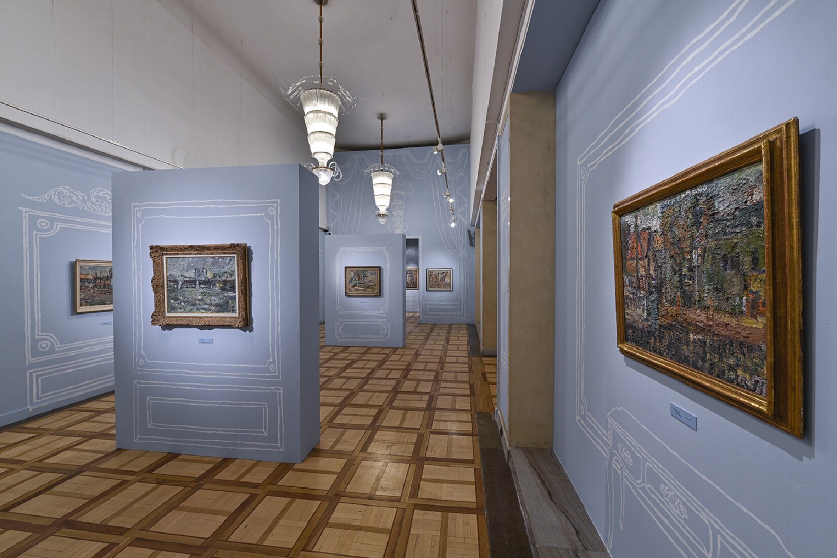 Zdjęcie przedstawia fragment wystawy we wnętrzu, gdzie na niebieskich ścianach są zawieszone obrazy w drewnianych ramach. Dodatkowo wokół każdego z nich na ścianie została narysowana dodatkowa kreskówkowa rama.