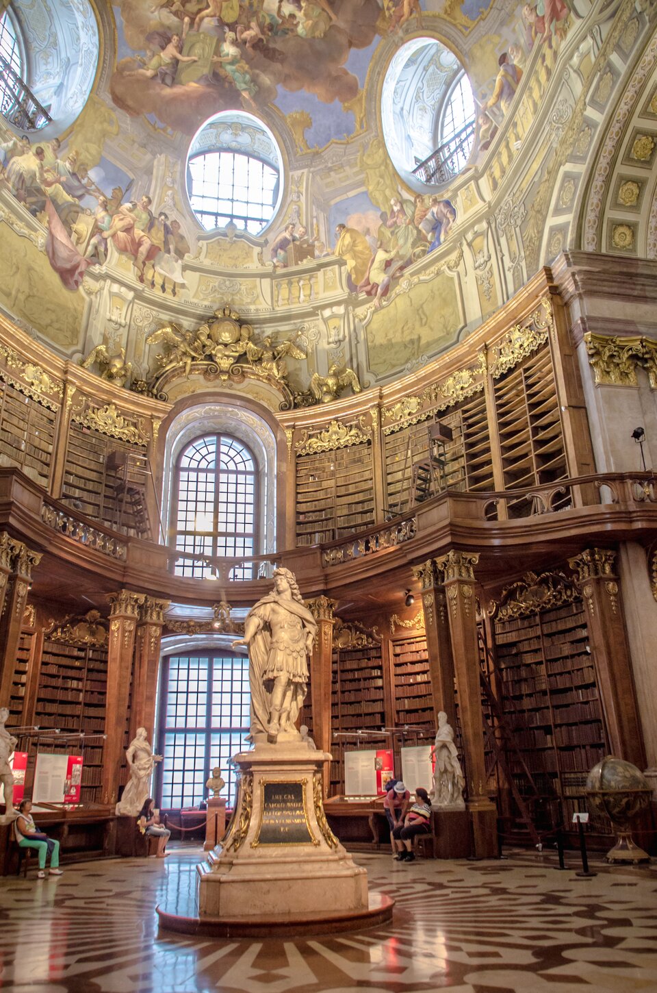 Ilustracja przedstawia wnętrze Biblioteki Dworskiej w Wiedniu, zaprojektowane przez Johanna Bernharda Fischer von Erlach i i Joseph Emmanuel Fischer von Erlach, a dokładnie salę paradną, serce Austriackiej Biblioteki Narodowej. Sala ma prawie 80 m długości i 20 m wysokości, zwieńczona jest kopułą, pięknie udekorowaną freskami autorstwa malarza dworskiego Daniela Grana. W kopule znajdują się okrągłe okna. Wnętrze stanowią drewniane półki, które są wypełnione rzędami książek po sam sufit. Przy regałach stoją drabiny. W połowie wysokości ścian znajduje się balkon opierający się na drewnianych kolumnach. Zarówno balkon, jak i kolumny są ozdobione złotym ornamentem. Półki na książki zdobią kartusze z ornamentami w stylu regencji, zawierające sygnatury. Na środku pomieszczenia, a także po bokach stoi rzeźby.