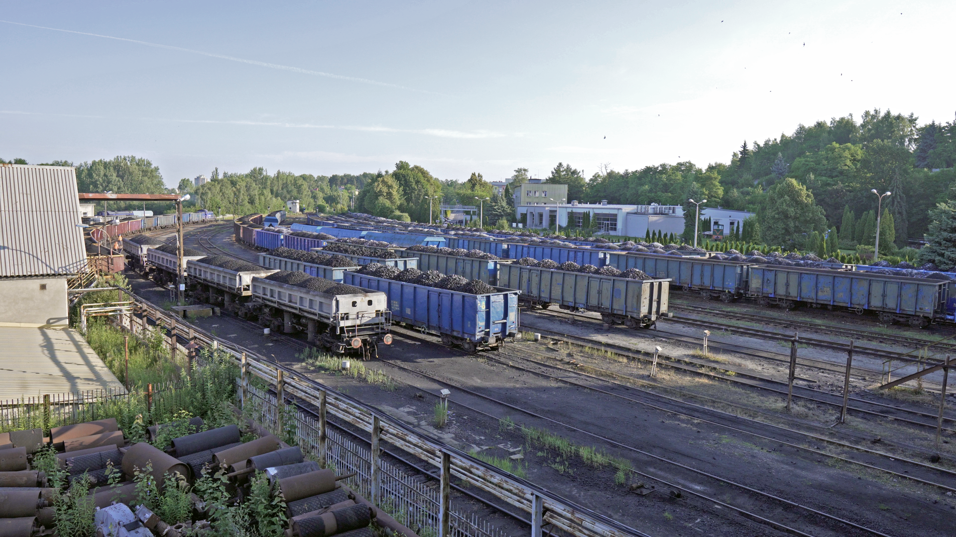 Na zdjęciu przedstawiono transport węgla przy pomocy kolei. Widoczne na zdjęciu wagony są załadowane węglem. Wagony stoją na szynach. Po lewej stronie zdjęcia znajduje się budynek kolejowy. Po prawej stronie niskie zabudowania oraz las.