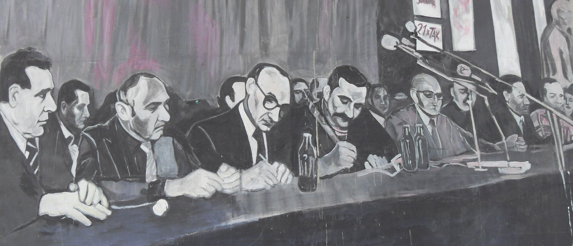 Zdjęcie przedstawia mężczyzn siedzących za stołem, podpisują oni dokumenty. Są ubrani w garnitury, mają skupione twarze. Za nimi siedzą inni ludzie. Ustawione są mikrofony, banery. 