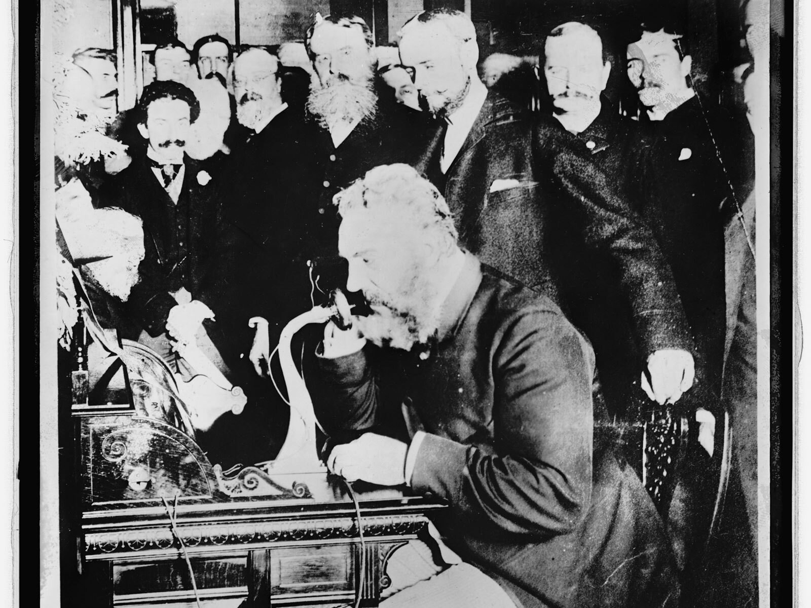 Czarno‑białe zdjęcie przedstawia mężczyznę siedzącego przy biurku, z którego wystaje długi mikrofon, podobny do rury, mężczyzna jest nachylony w jego kierunku. Wokół niego stoi duża grupa innych mężczyzn przyglądających się osobie przy biurku.