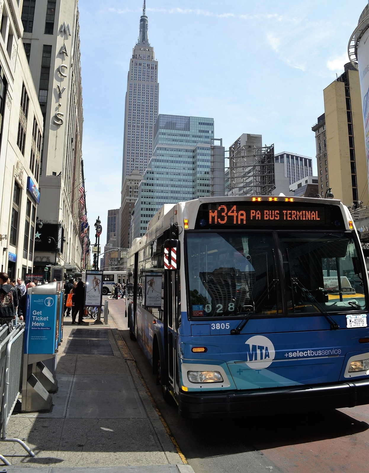 Ilustracja przedstawia autobus komunikacji miejskiej, który zatrzymał się na przystanku. Autobus jest w kolorze niebieskim. Jego oznaczenie linii to numer M34A - PA BUS TERNIMAL. W oddali widoczne są budynki miejskie.