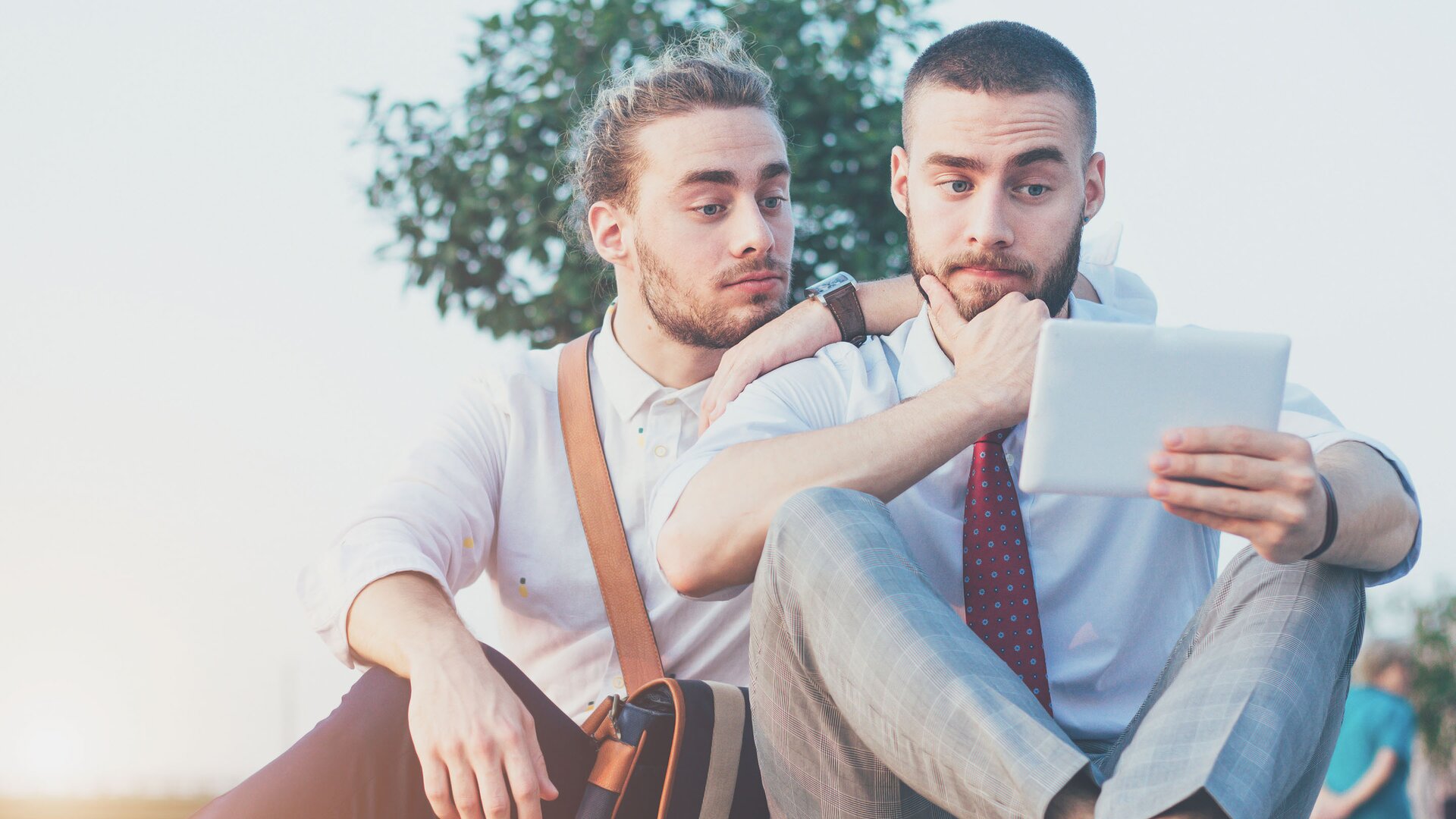 Zdjęcie przedstawia braci bliźniaków, którzy wyglądają podobnie. Obaj mają brodę. Ich fryzury różnią się: jeden z nich ma długie, upięte włosy, a drugi krótkie. Bracia wpatrują się w tablet.