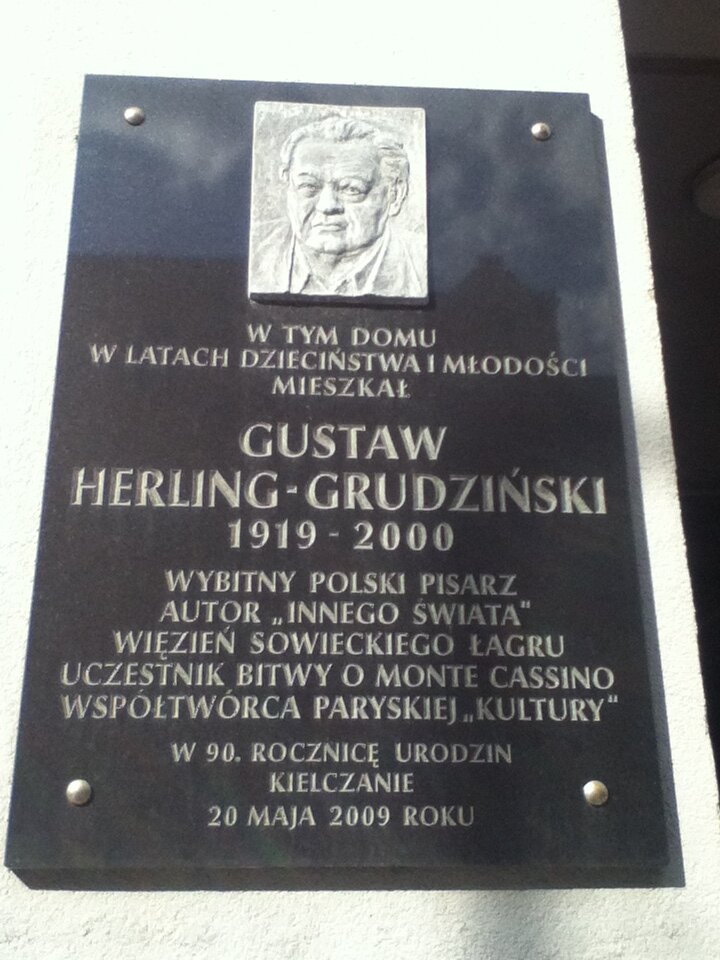 Zdjęcie przedstawia tablicę pamiątkową. U góry tablicy znajduje się portret dojrzałego mężczyzny. Jest to popiersie ukazujące całą, kwadratową twarz pisarza. Postać ma lekkie zakola. Czoło mężczyzny jest wysokie, zmarszczone. Ma przerzedzone brwi i wąskie oczy z opuchniętymi powiekami. Nos mężczyzny jest szeroki, a usta skrzywione. Pisarz ma dołek na podbródku. Jest ubrany w koszulę. Pod spodem umieszczono napis: W tym domu w latach dzieciństwa i młodości mieszkał Gustaw Herling‑Grudziński, od 1919 do 2000, wybitny polski pisarz, autor Innego świata, więzień sowieckiego łagru, uczestnik bitwy o Monte Cassino, współtwórca paryskiej Kultury. W dziewięćdziesiątą rocznicę urodzin kielczanie, 20 maja 2009.