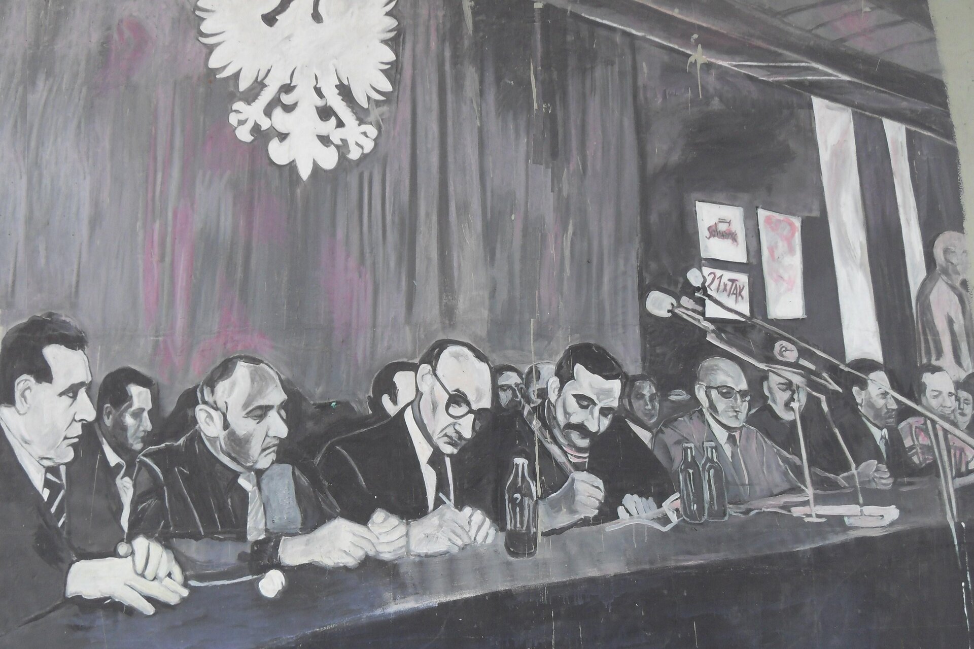 Rysunek przedstawia mężczyzn siedzących za długim stołem. Dwóch po środku podpisuje dokumenty. Mężczyzna z wąsem trzyma większy długopis. Za nimi pozostali mężczyźni, którzy siedzą. Za nimi na ścianie orzeł. 