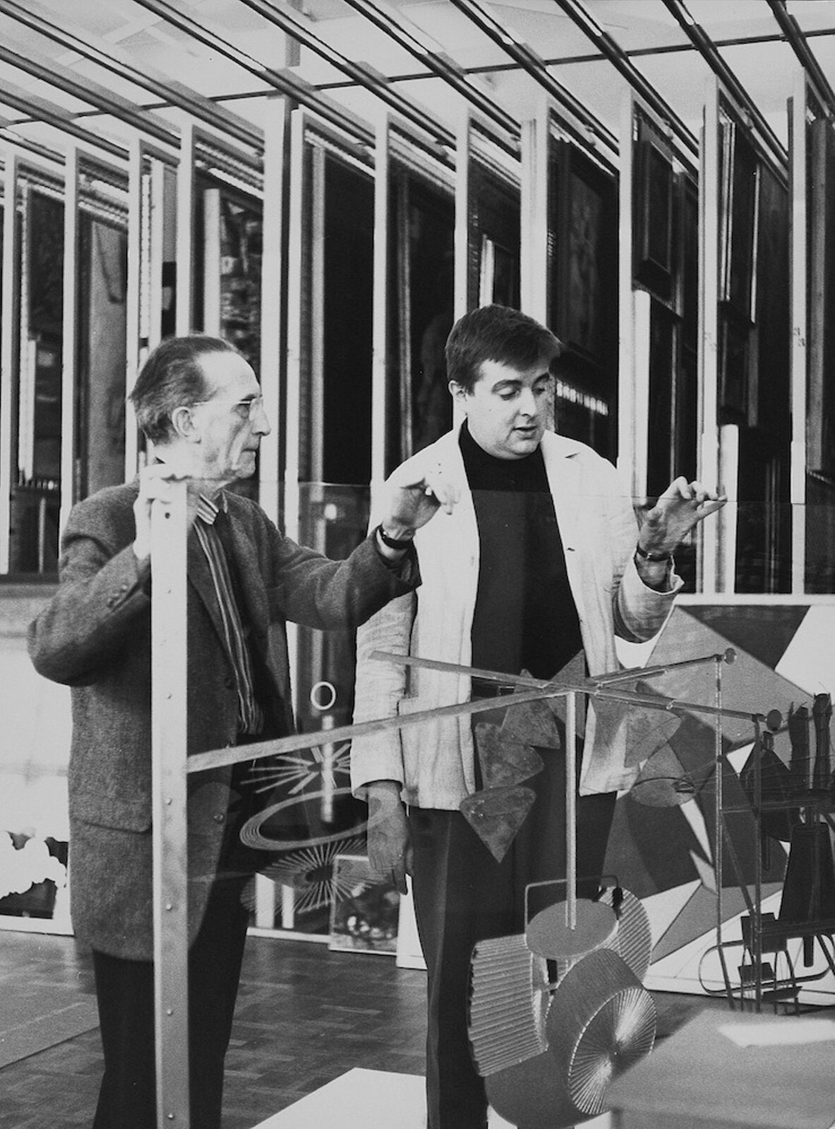  Fotografia przedstawia Marcela Duchampa [czytaj: marsela duszą] oraz Ulfa Linde przed instalacją artystyczną. Mężczyźni stoją przed szybą, Duchamp [czytaj: duszą] pokazuje elementy pracy. Za wielką szybą widoczne są różnego rodzaju rzeczy zawieszone na pałąku – między innymi przedmioty przypominające patelnię i miski.