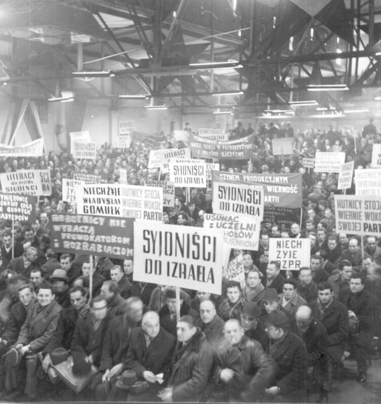 Zdjęcie przedstawia halę fabryczną Siedzą w niej zgromadzeni robotnicy, Trzymają transparenty z napisami: Syjoniści do Izraela, Robotnicy nie wybaczą prowokatorom i rozrabiaczom, Niech żyje PZPR, Niech żyje Władysław Gomułka, Hutnicy stoją wiernie wokół swojej Partii.