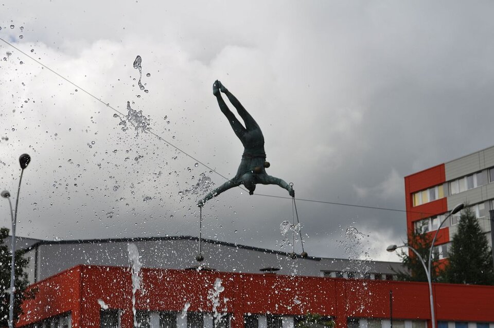 Ilustracja przedstawia balansującą, wiszącą na linie rzeźbę Jerzego Jotki Kędziory, znajdującą się w Częstochowie. Dzieło przedstawia akrobatę wykonującego sztuczki. Stoi on na głowie na linie. Z obu rąk zwisa mu po parze kulek na sznurkach. Fotografia jest wykonana od dołu, lekko z boku -rzeźba wydaje się znajdować powyżej zabudowań. Dodatkowo w kadrze jest górna cześć tryskającej wody z fontann. 