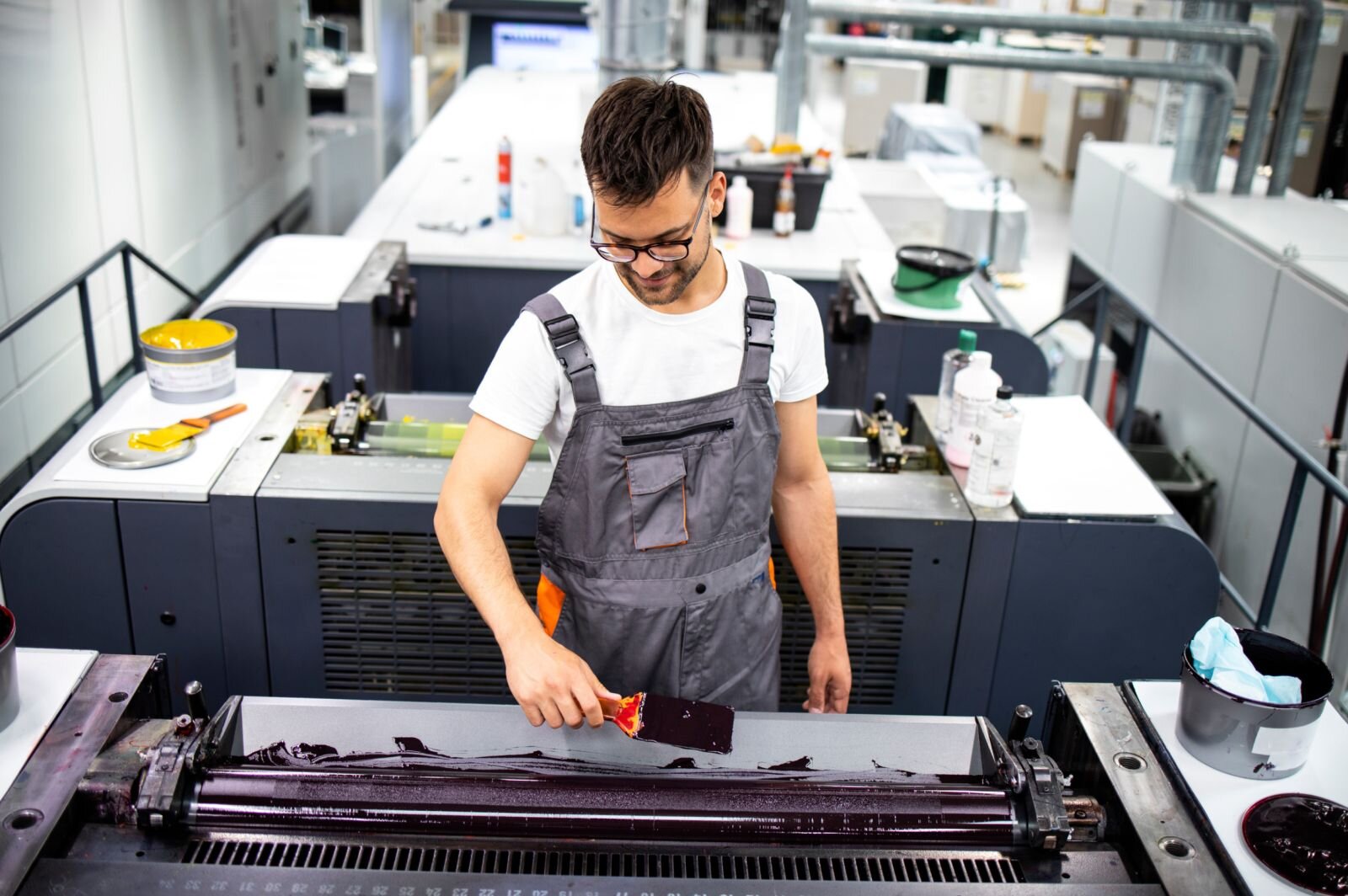 Zdjęcie ukazuje pracownika drukarni w stroju roboczym przed ploterem. W pomieszczeniu z tyłu stoją jeszcze dwa podobne urządzenia. Ploter drukujący jest połączony z komputerem i może drukować cyfrowo duże formaty na różnych powierzchniach. To nowoczesne urządzenie wykorzystywane w grafice użytkowej, do drukowania plakatów, bilbordów i banerów.