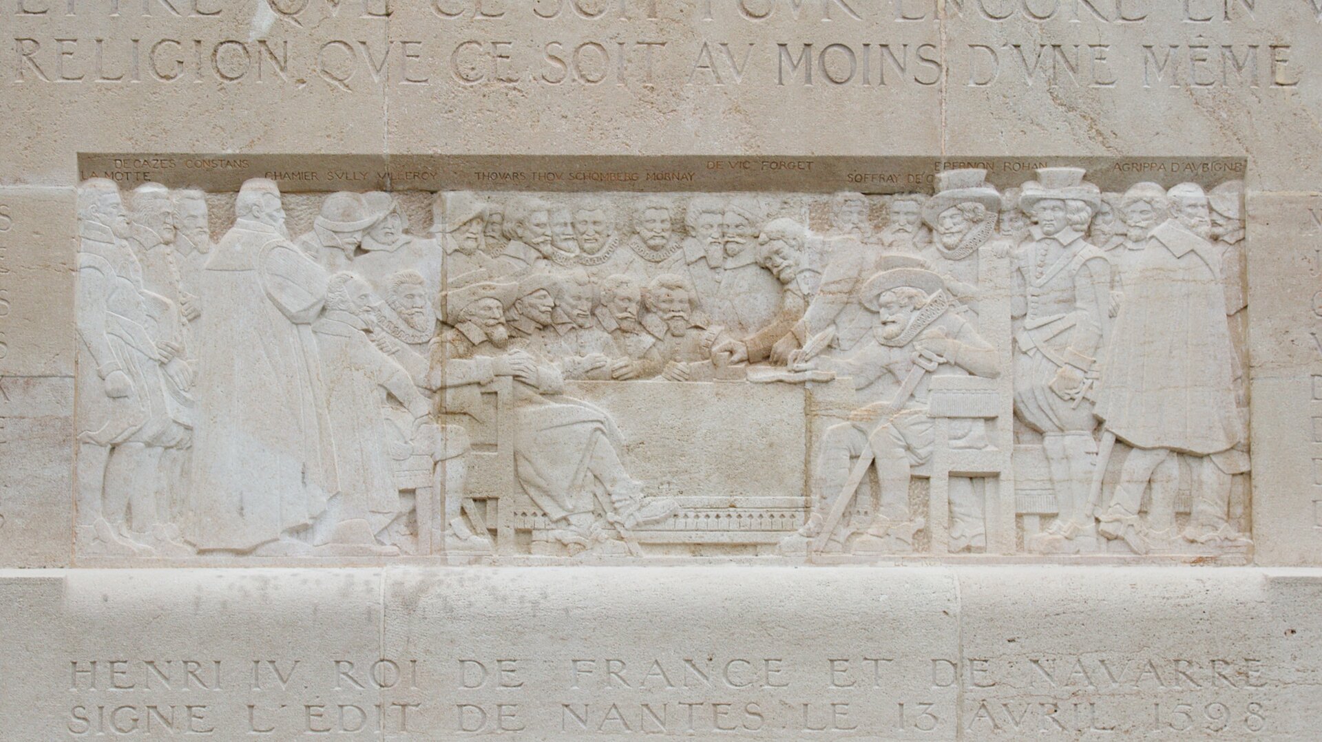 Na zdjęciu płaskorzeźba. W środku przy stole siedzą mężczyźni. Wszyscy mają brody i wąsy. Po prawej stronie siedzi mężczyzna z piórem w czapce. Obok niego schyla się do stołu mężczyzna, który stawia na stole kubek. Z lewej i prawej strony oraz za siedzącymi przy stole stoją i przyglądają się siedzącym inni mężczyźni. Nad i pod przedstawioną sceną wyrzeźbione napisy w języku obcym