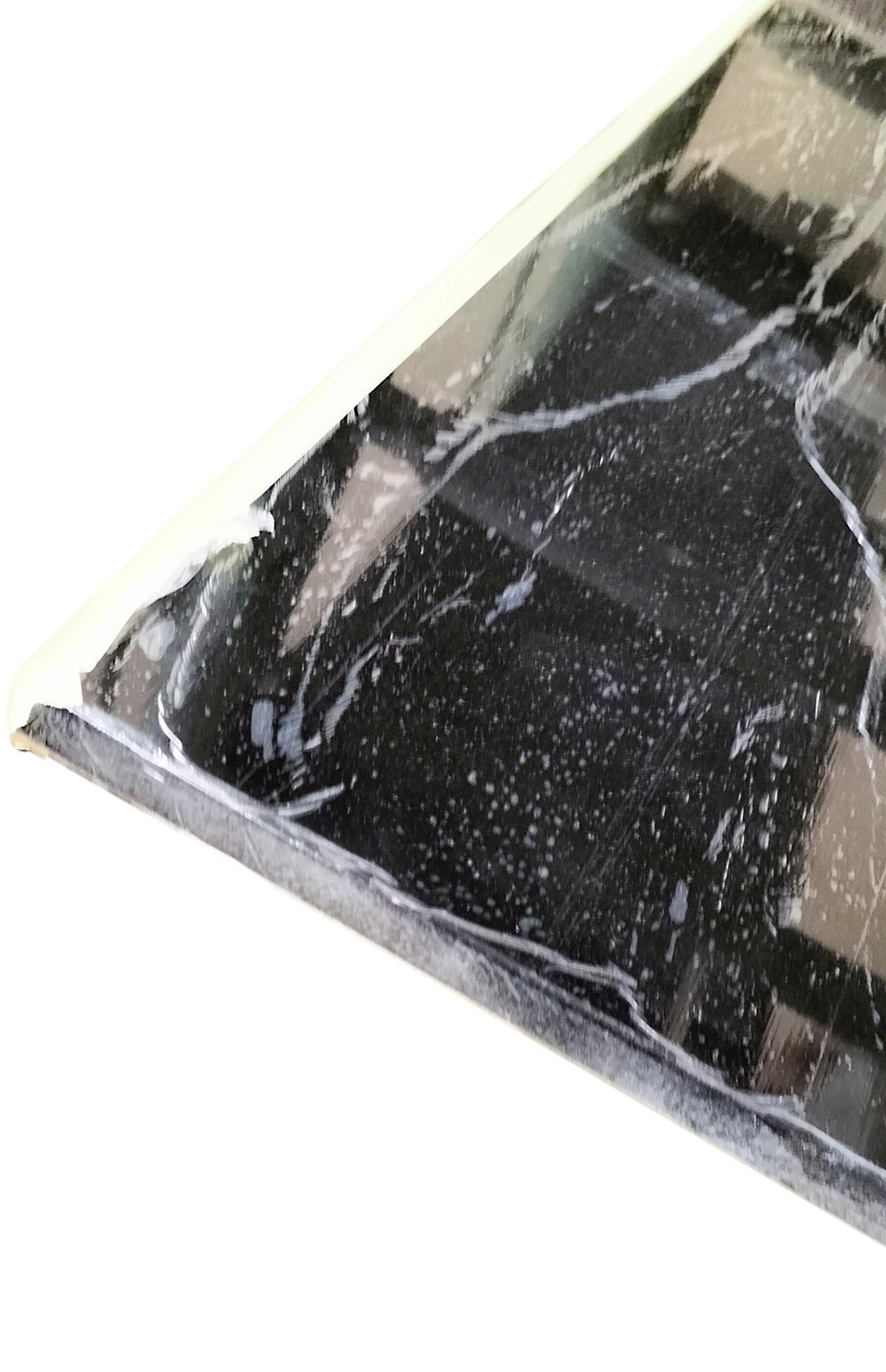 Zdjęcie przedstawia element obudowy pianinowej polakierowany czarnym lakierem poliestrowym, wypolerowany i owinięty folią strecz. Widoczny jest fragment prostokątnego elementu w kolorze szarym z kwadratami i prostokątami w kolorze czarnym. Przy lewej górnej krawędzi widoczny jest jasno szaro‑żółty pas symbolizujący taśmę klejącą.