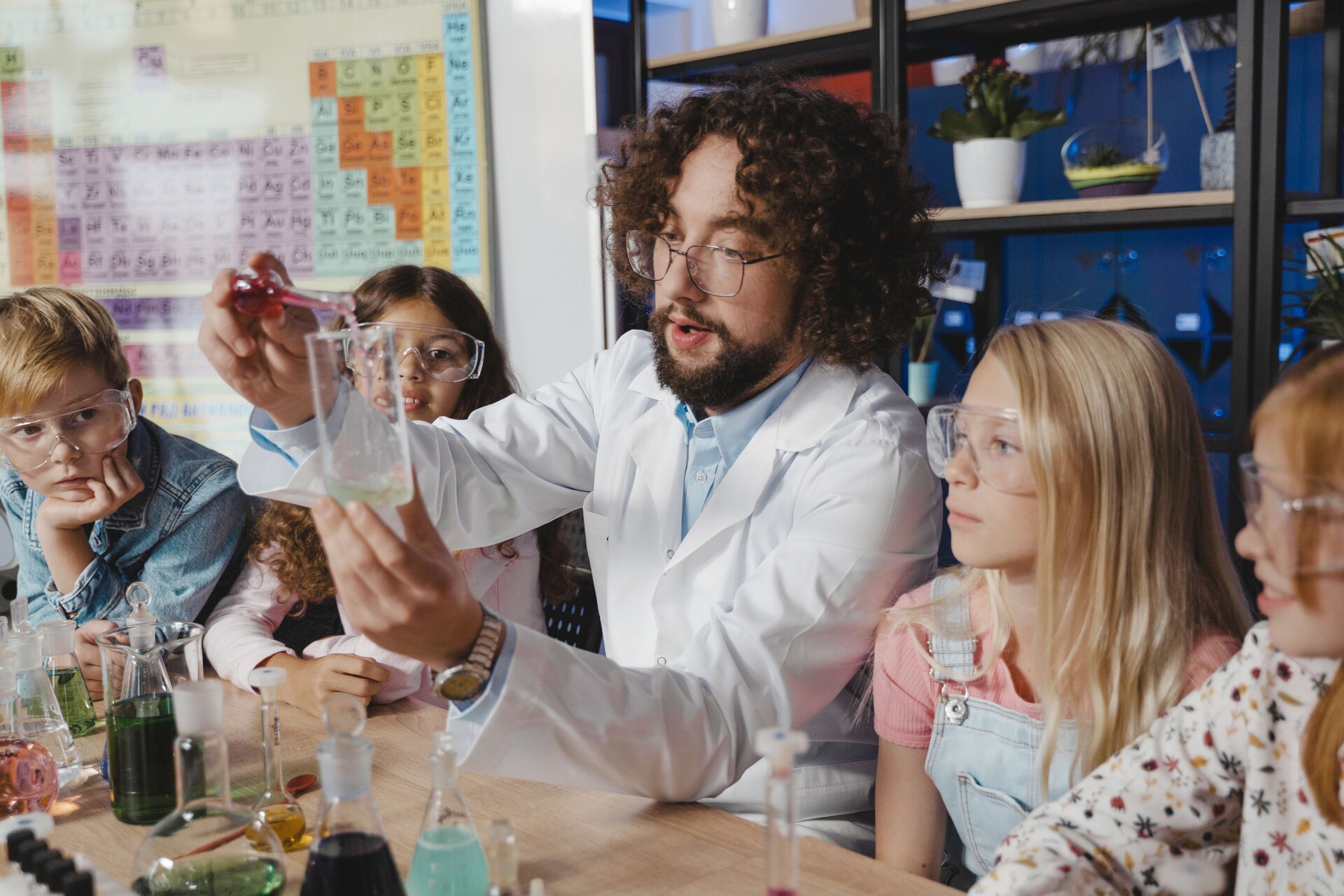 Ilustracja przedstawia nauczyciela chemii oraz czwórkę dzieci. Nauczyciel wykonuje eksperyment w klasie chemicznej. Dzieci w skupieniu i z uwagą obserwują, co się dzieje na zajęciach. W tle widać zlewki, kolby i inne sprzęty niezbędne do lekcji chemii.