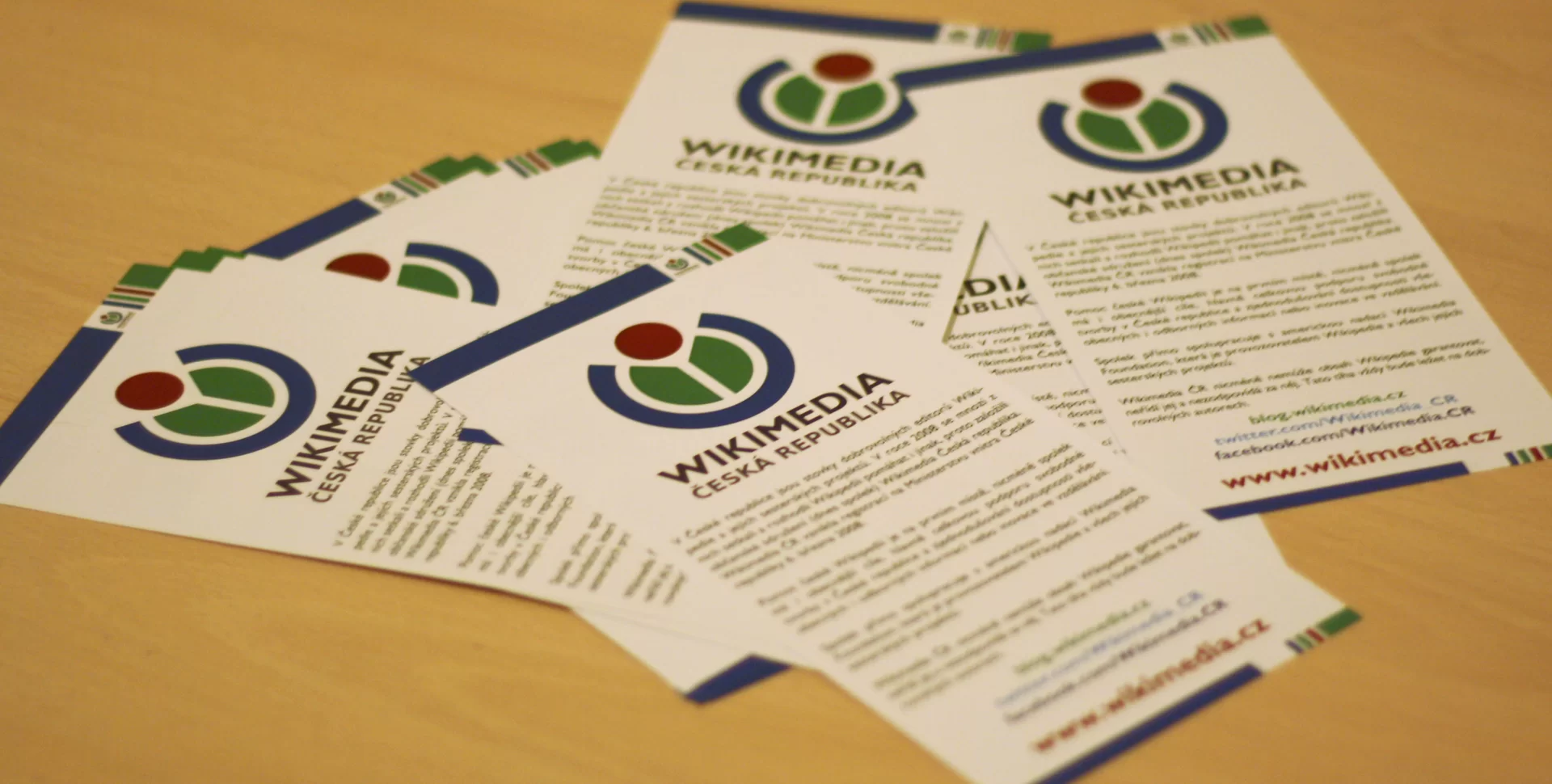 Ulotki leżące na stole, należące do Czeskiego oddziału Wikimedia. Ulotka posiada na środku u góry logo Wikimedia - granatowy niezamknięty od góry gruby okrąg, w którego środku znajdują się dwie zielone równe wydzielone części koła. Granatowy okrąg domyka bordowa kula, jednak elementy loga sie nie stykają. Ulotki zawierają 4 akapity różnej długości oraz linki prowadzące do czeskich stron Wikimedia.