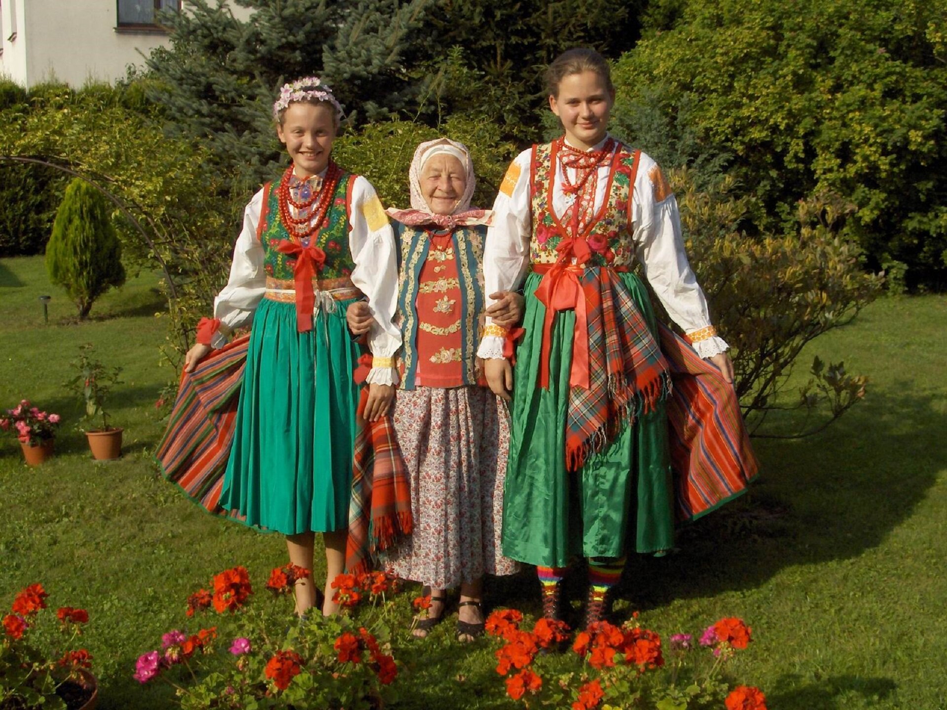 Zdjęcie przedstawia wilamowski strój ludowy. Na fotografii ukazane są dwie młode dziewczyny oraz starsza kobieta. Strój charakteryzuje się zielone spódnice, białe koszule oraz chusty. Kobiety mają na szyi czerwone korale. Starsza kobieta na głowie ubraną ma chustę. Jedna z dziewczyn ubrany ma wianek.