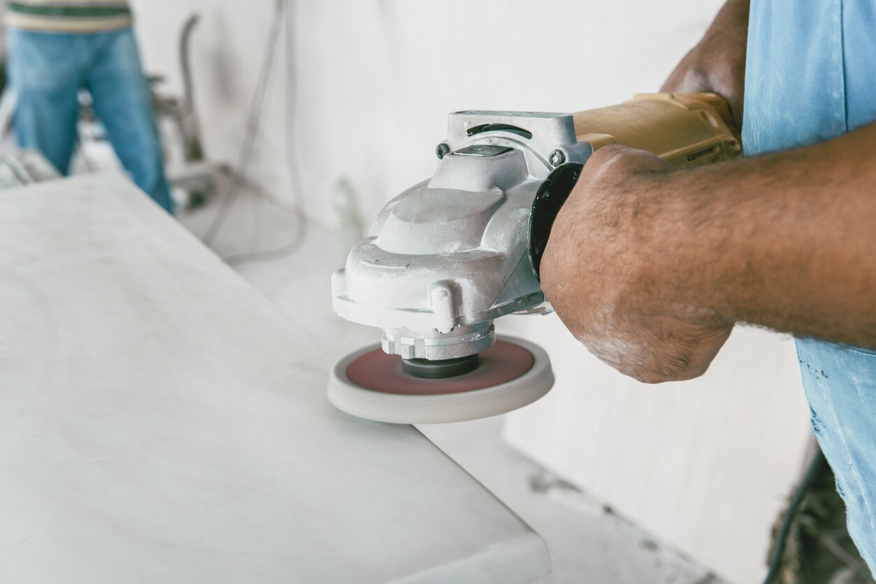 Zdjęcie przedstawia marmurową płytę. Jej brzeg jest szlifowany przez kamieniarza, który trzyma w obydwu rękach specjalistyczne narzędzie przeznaczone do tej czynności.