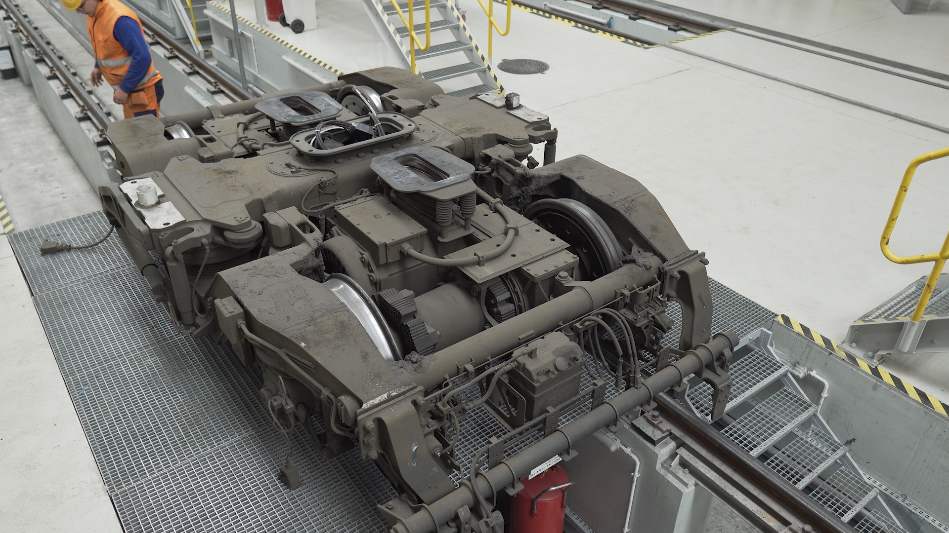 Ilustracja przedstawia wózek jezdny w widoku z góry, wózek znajduje się na kanale warsztatowym, konstrukcją przypomina prostokątną ramę ze środkowym elementem poprzecznym, w którym umieszczony jest przegub skrętny. Posiada dwie pary kół, które stykają się z szynami, na których jest usadzony. Wózek jezdny to element nadwozia pojazdu szynowego.